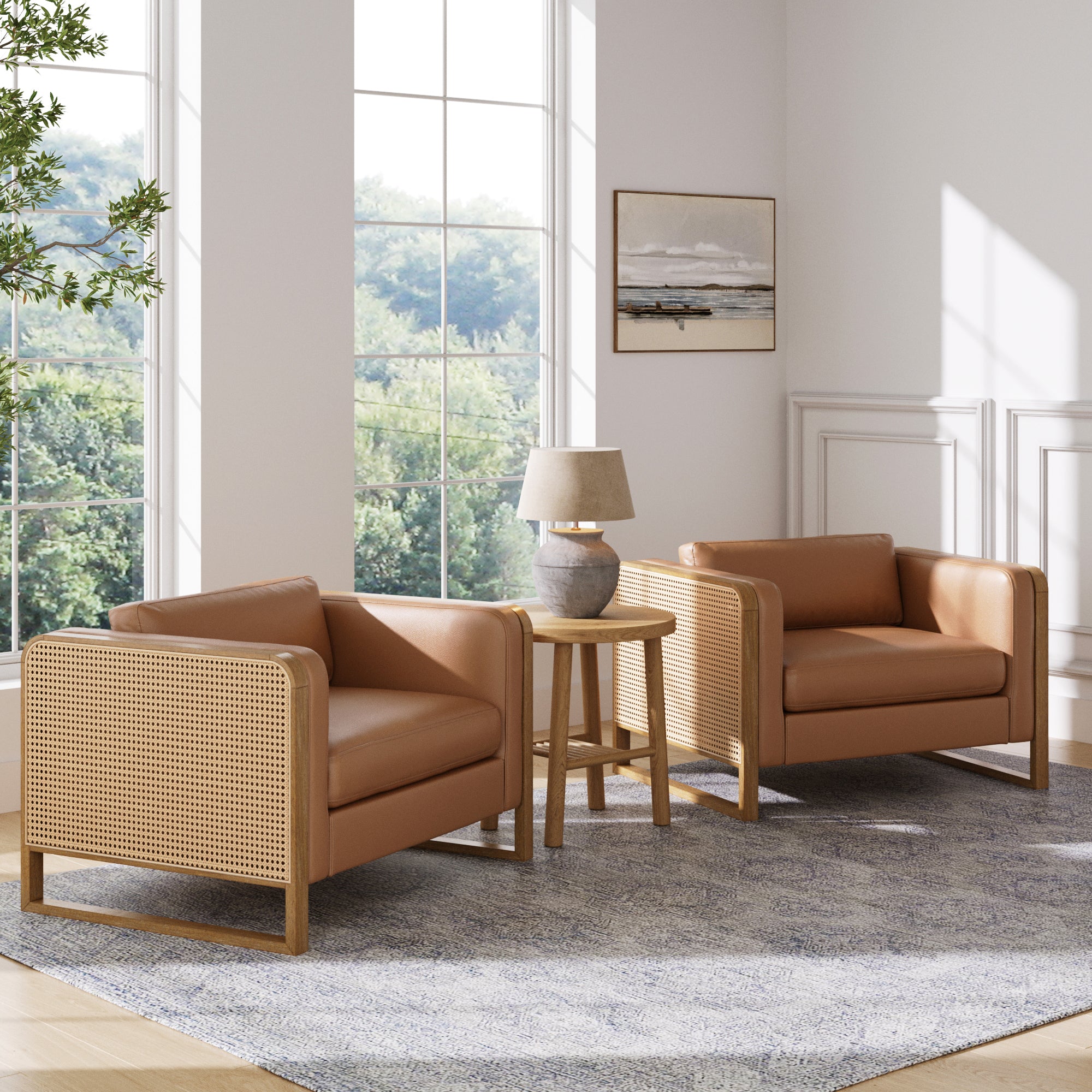 Two Nathan James Solid Wood Accent Chairs with rattan arms in camel are placed on a gray rug by large windows. A small round table with a lamp sits between the chairs, and a framed picture hangs on the wall. Trees can be seen outside.