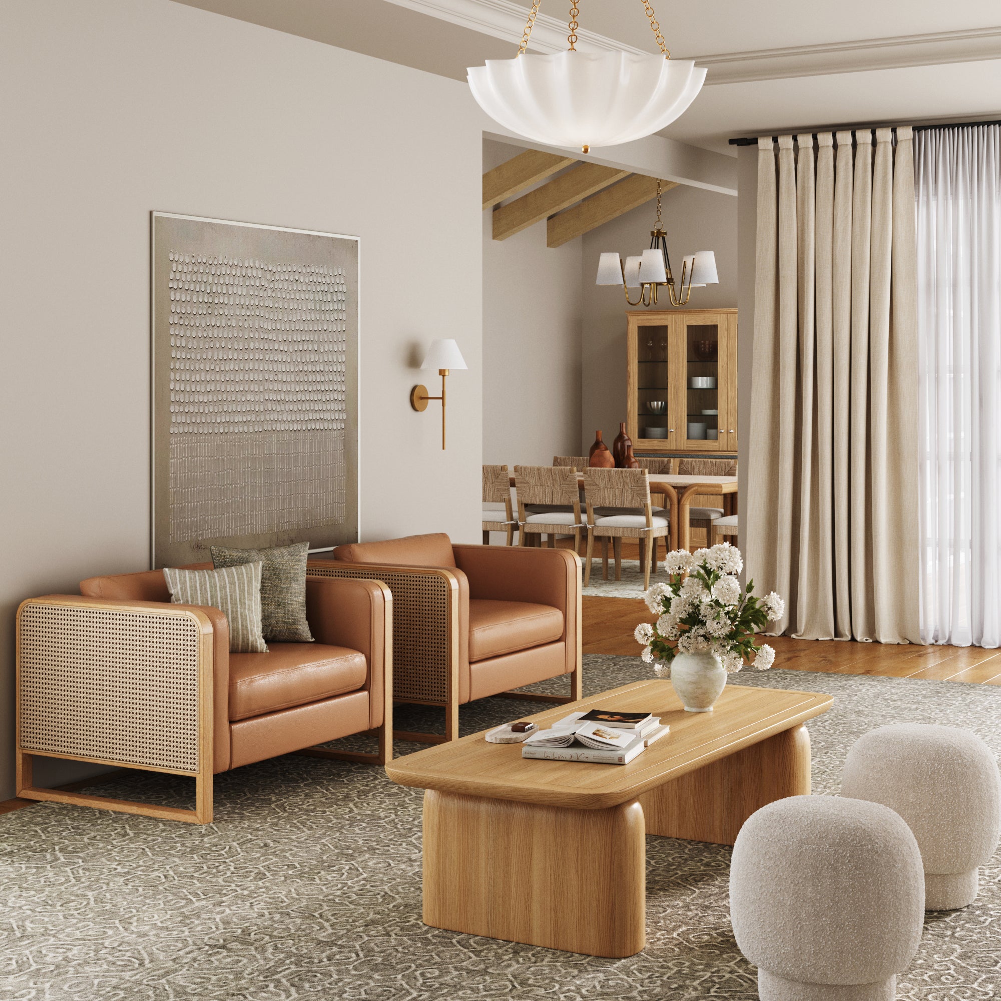 A modern living room with tan leather armchairs, a wooden coffee table with a vase of flowers, two round ottomans, neutral decor, large windows with beige curtains, and a dining area in the background.
