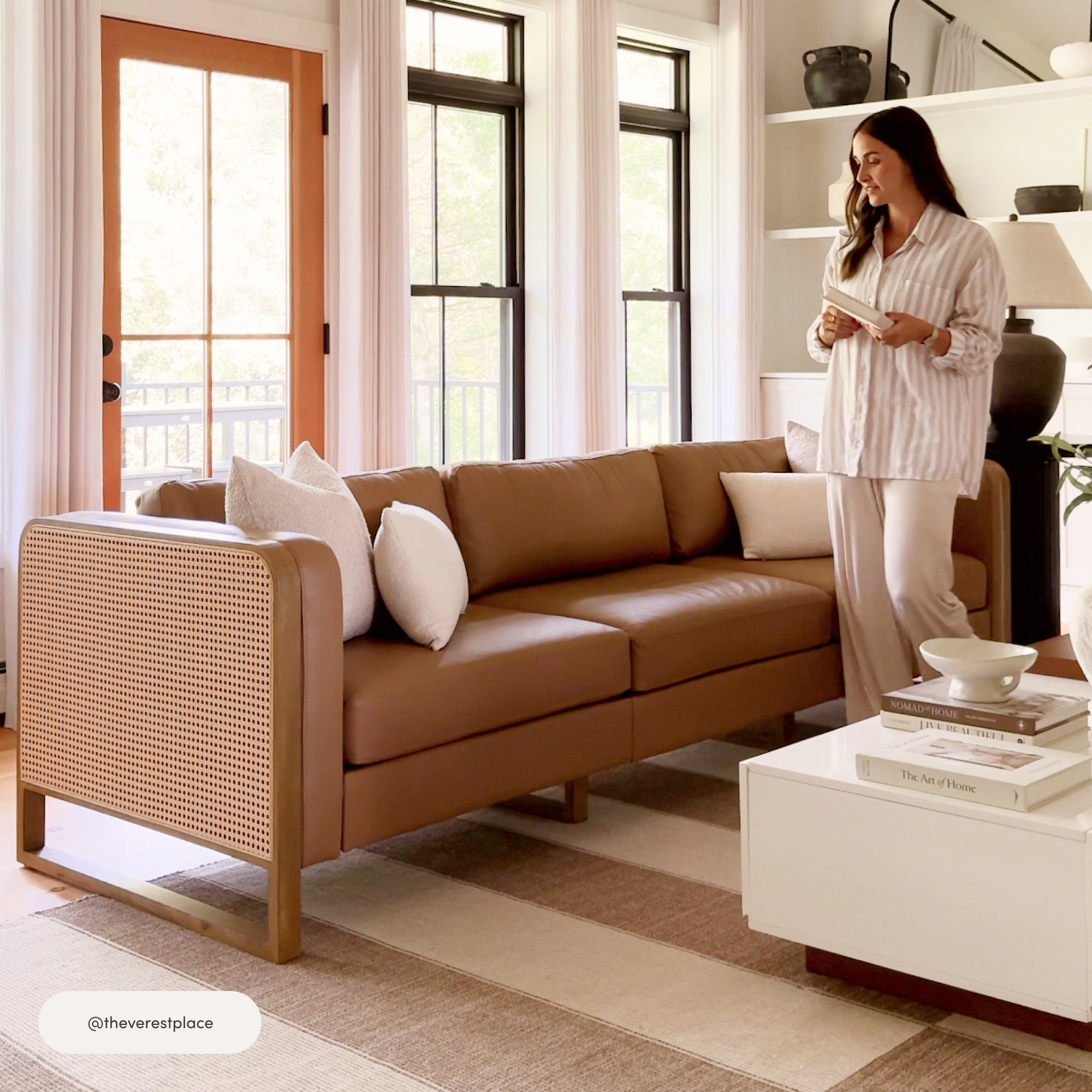 A woman in pajamas stands in a bright living room holding a book, next to a tan leather and cane sofa with white cushions. The space features large windows, a white coffee table, and minimalist decor.