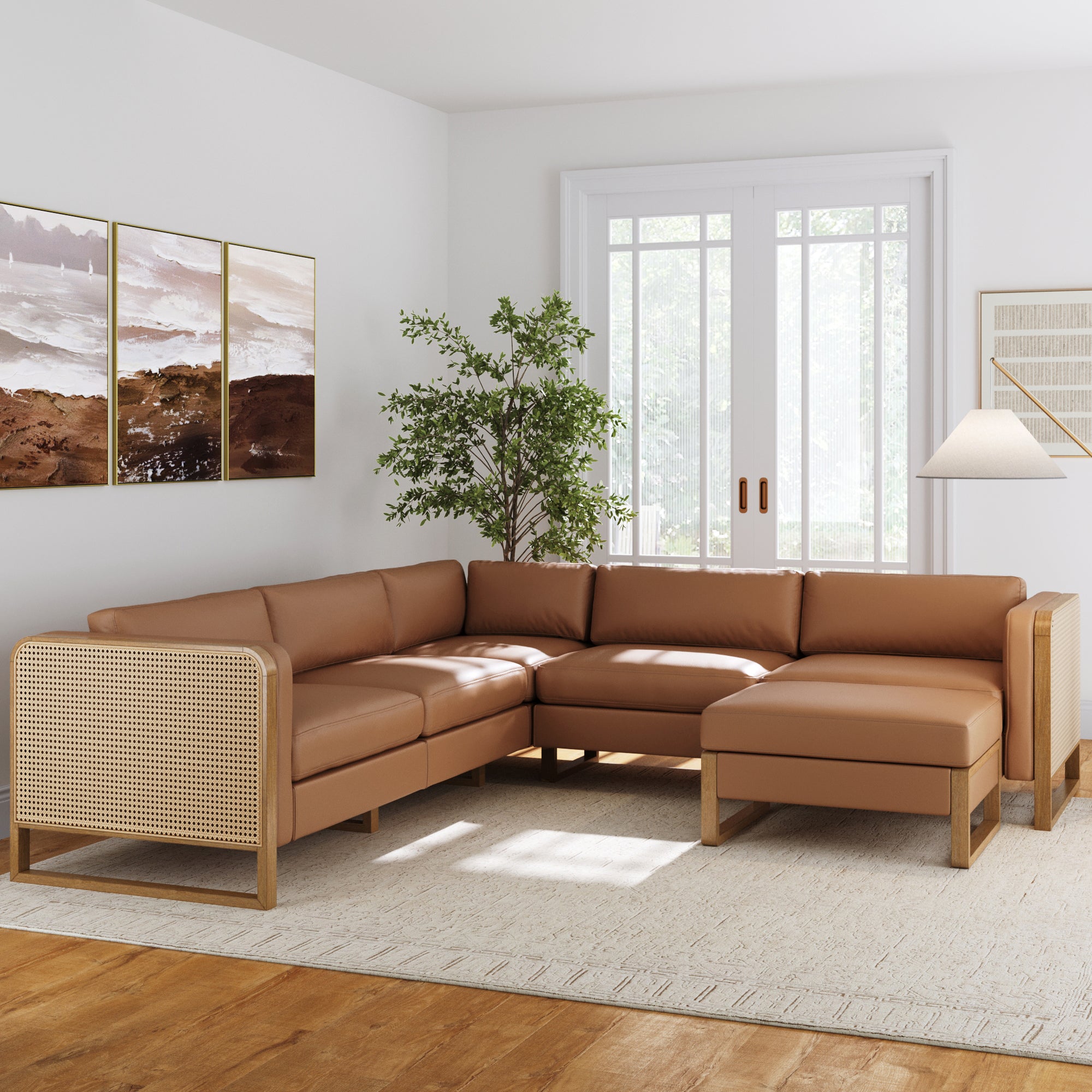 A modern living room features the Nathan James Rattan 5-Seat Sectional with Ottoman Set Wood Frame Camel, a large window, light walls, a beige rug, landscape wall art, a green plant, and a standing lamp.