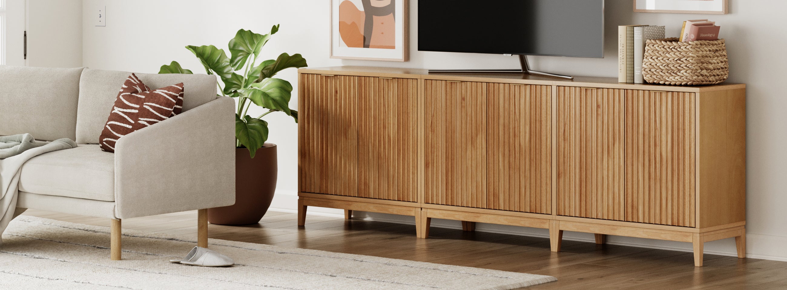 A modern living room with a light-colored sofa, a patterned pillow, a wooden cabinet, a potted plant, a TV, baskets, and abstract wall art, all on a light rug and wooden floor.
