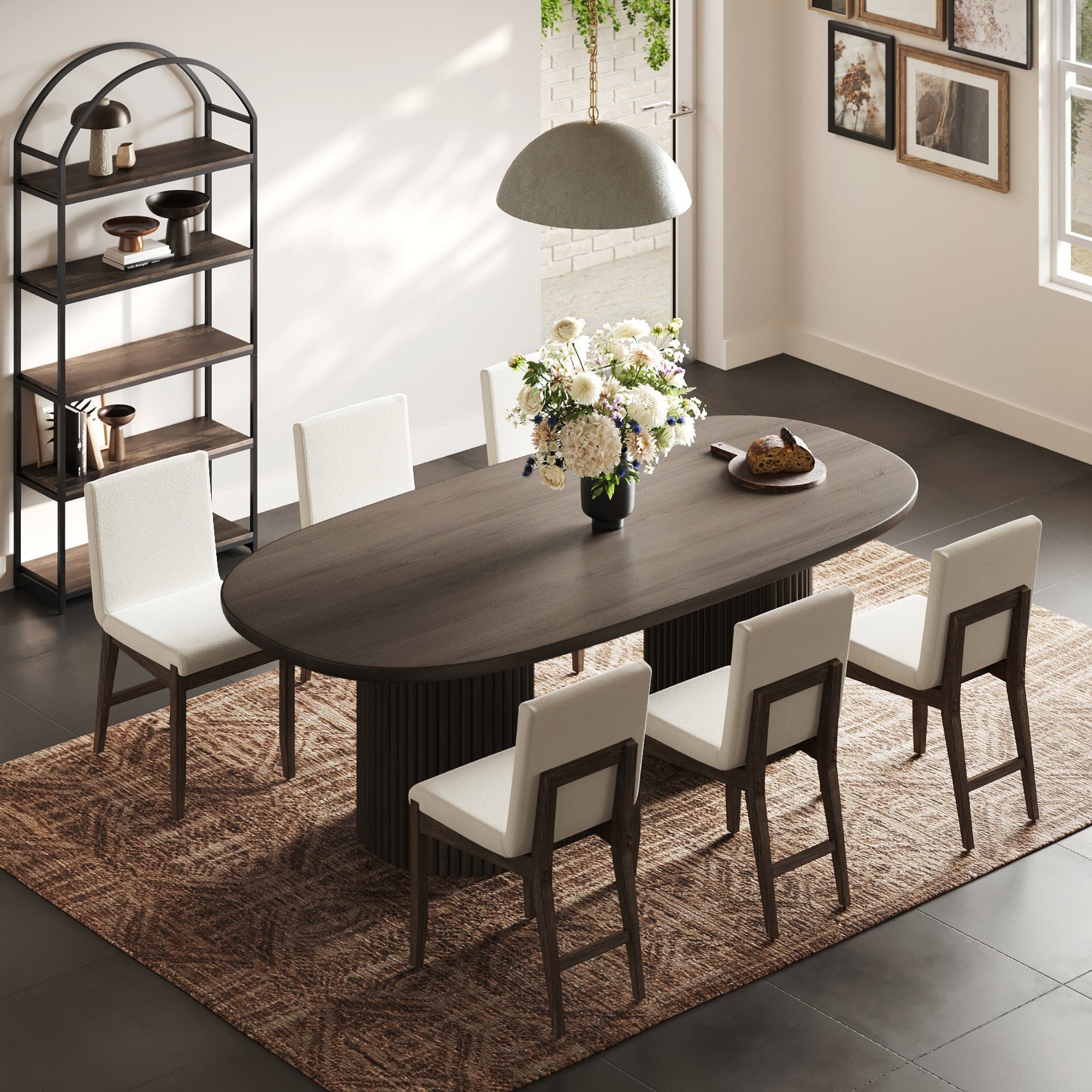 A modern dining room features a dark solid wood oval table, six Nathan James Exposed Back Dining Chairs in dark brown, a large floral centerpiece, hanging lamp, open shelving, framed wall art, and a patterned rug on tiled floors lit by sunlight.