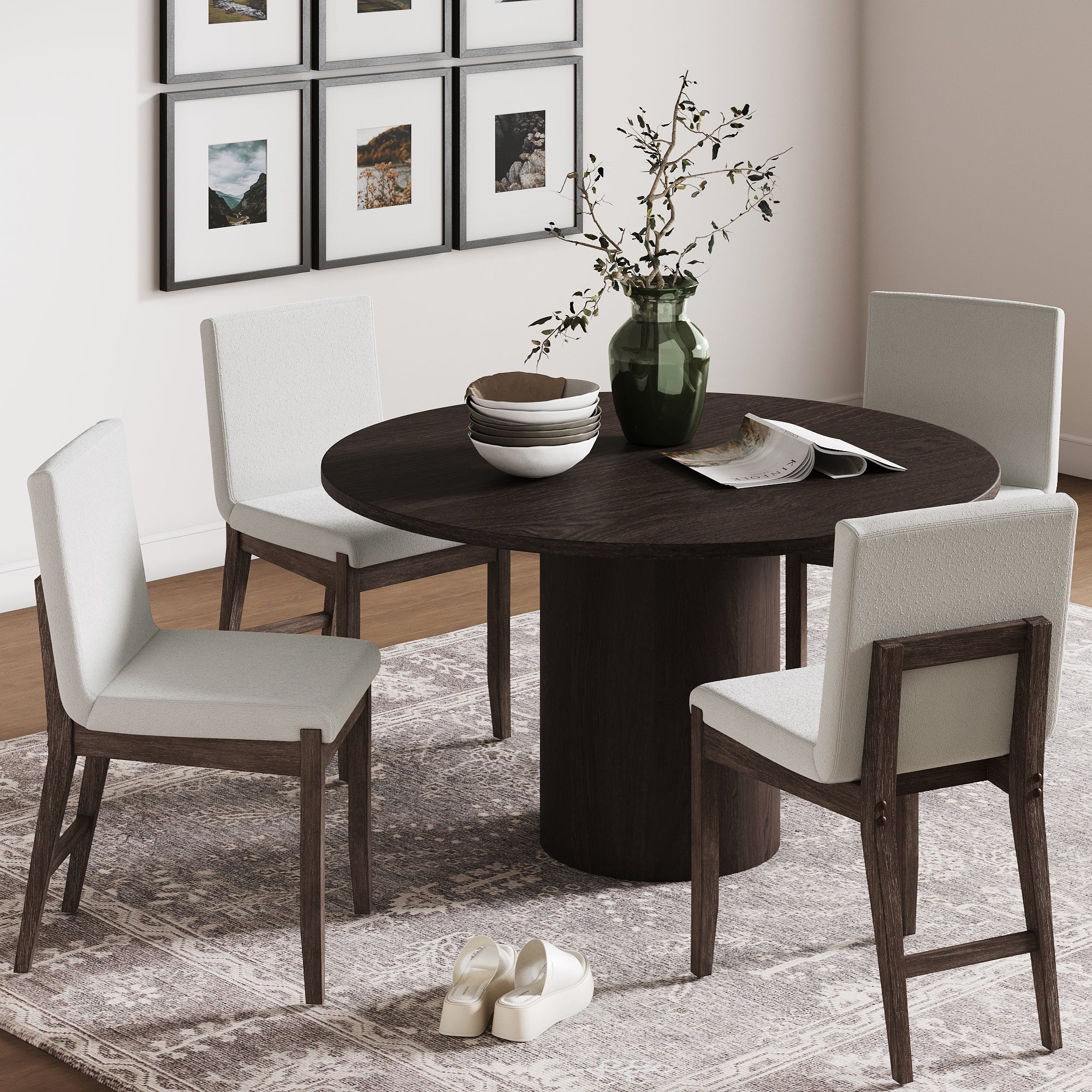 A round dark wood dining table with four Nathan James Exposed Back Dining Chairs in dark brown boucle sits on a patterned rug. The table holds a green vase, bowls, and a magazine, while white slippers and framed art complete the space.