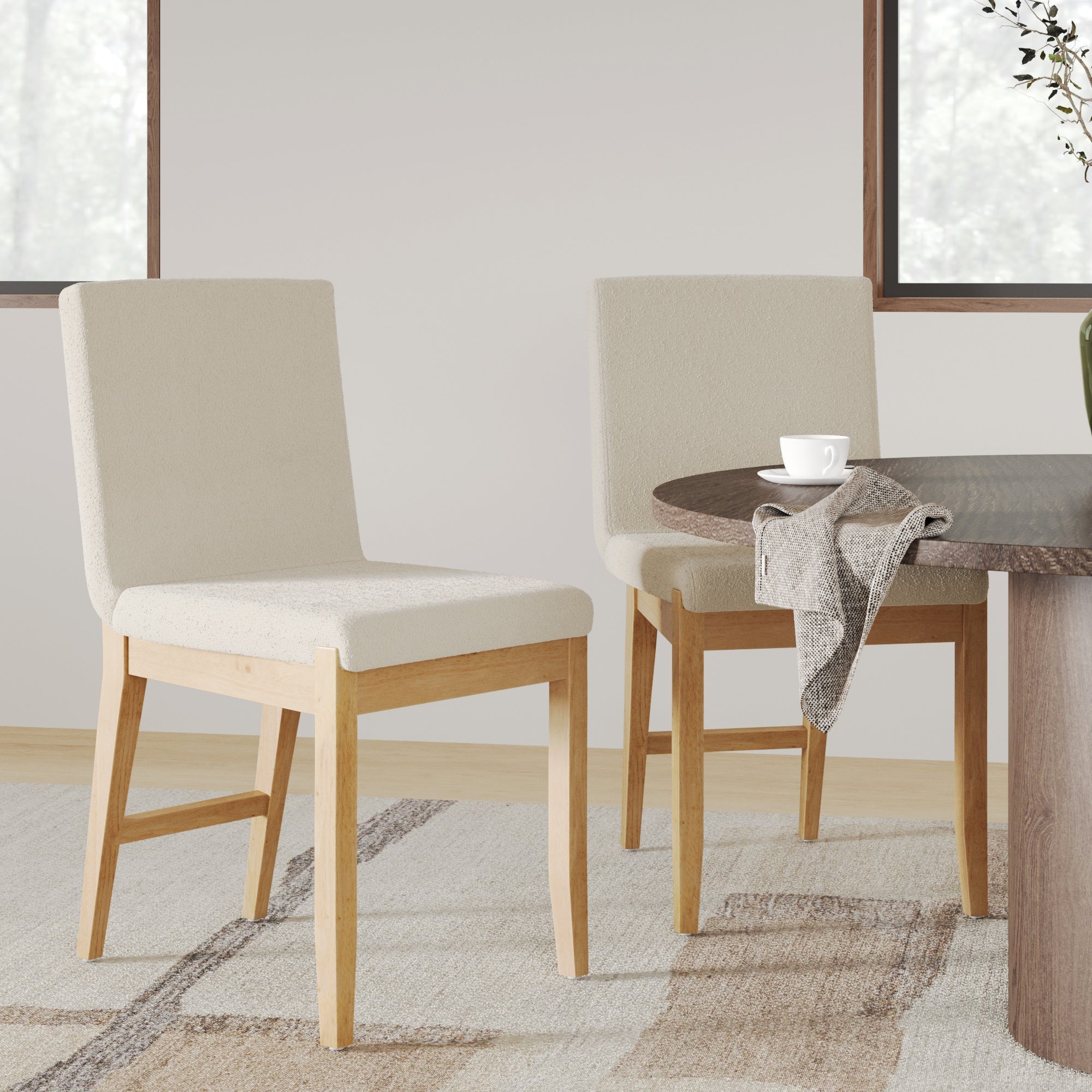 A set of two Nathan James Modern Exposed Back Dining Chairs in beige sits by a round wooden table with a cup, saucer, and napkin. The light-walled room features a patterned rug and large windows in the background.