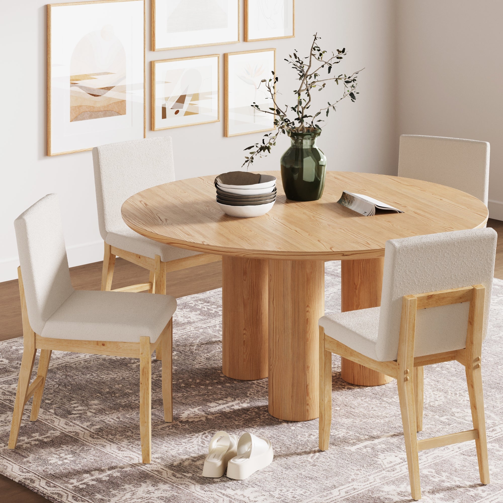 A round wooden dining table with four Nathan James Exposed Back Dining Chairs in Brushed Warm Pine sits on a patterned rug. A green vase with branches, plates, and an open book are on the table; art prints hang above and slippers rest nearby.