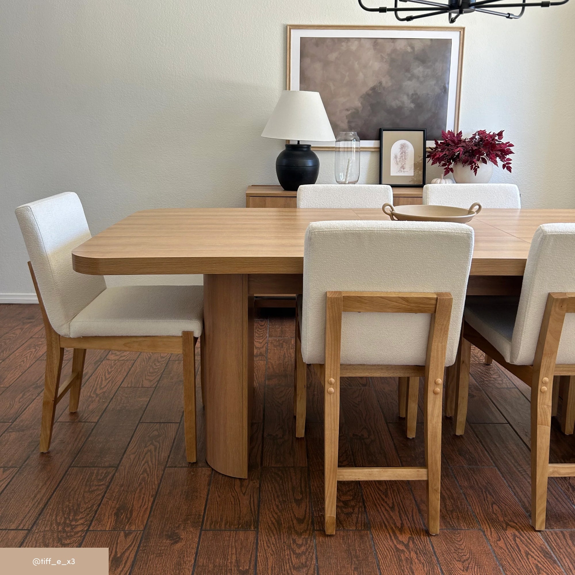 A modern dining room with a wooden table, six cream-upholstered chairs, a sideboard, a table lamp, framed artwork, and a vase with red foliage, all set on a wood floor.