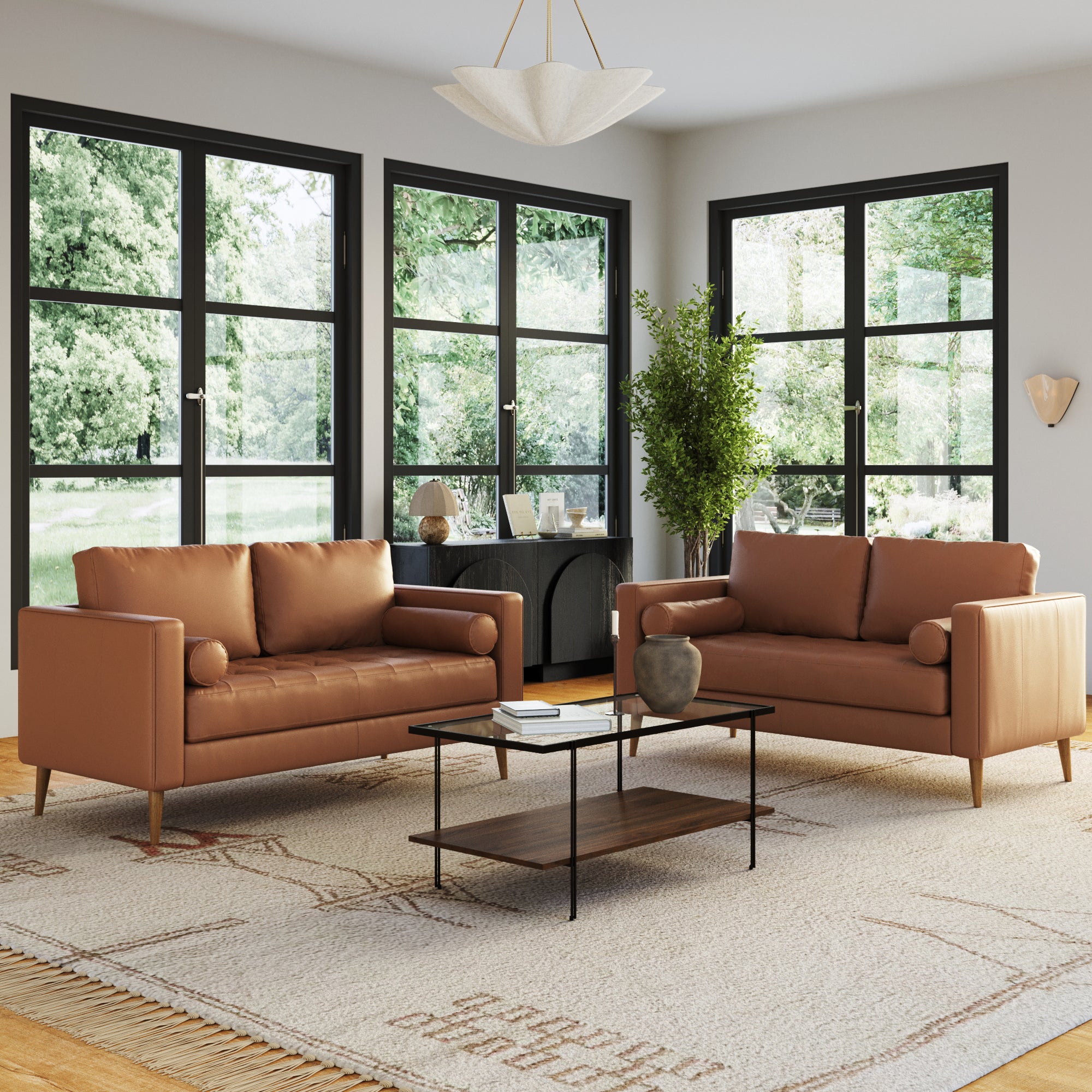 A modern living room featuring the Nathan James Mid-Century Tufted Leather Loveseat Sofa with Bench Seat in camel, a glass coffee table, large black-framed windows, geometric rug, plant, sideboard, and lush greenery outside.
