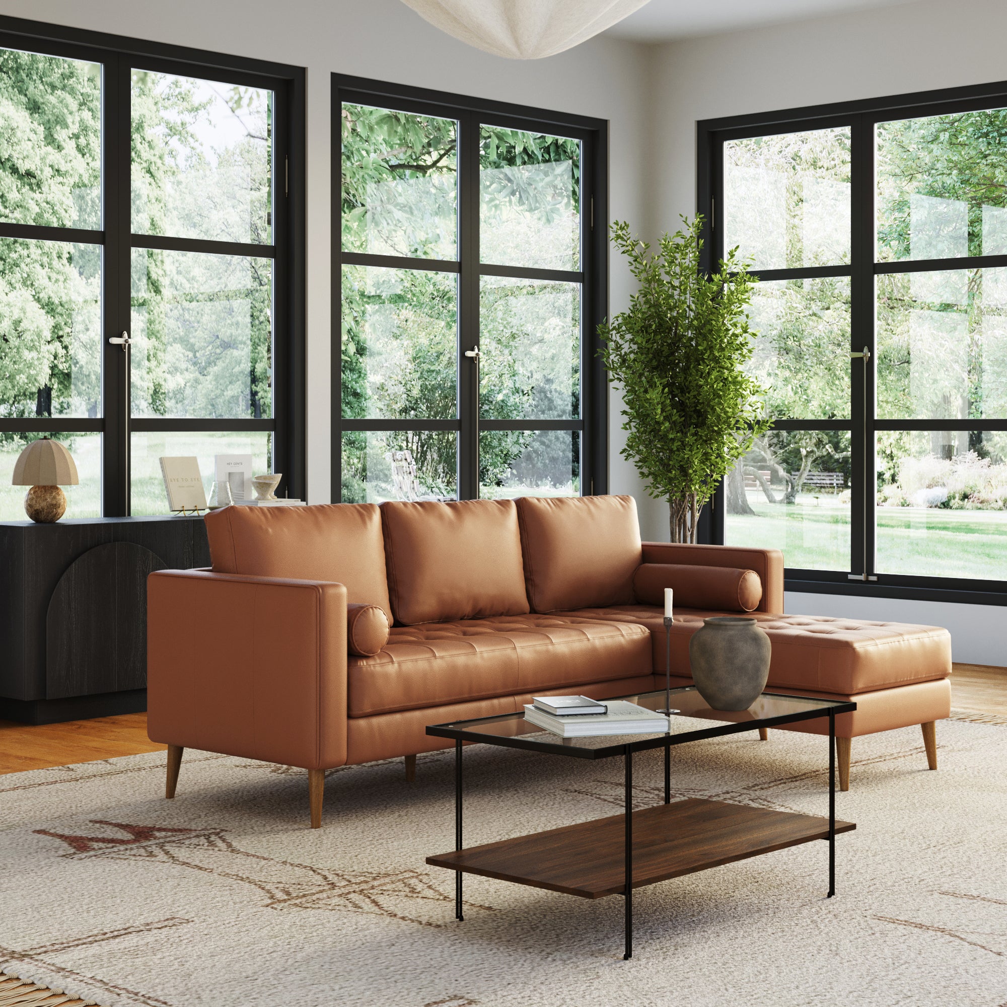A modern living room with large windows features the Nathan James Mid-Century Tufted Leather 3-Seat Sofa with Bench Seat & Chaise in camel, a black and wood coffee table, a potted tree, and a light rug on wooden floors filled with natural light.
