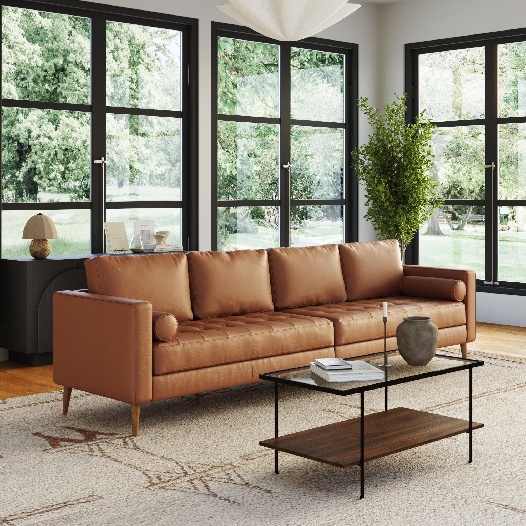 A modern living room with the Nathan James Mid-Century Tufted Leather 4-Seat Sofa with Bench Seats in camel, a glass coffee table with a wooden shelf, a potted plant, small decor desk, large windows, and views of lush greenery.