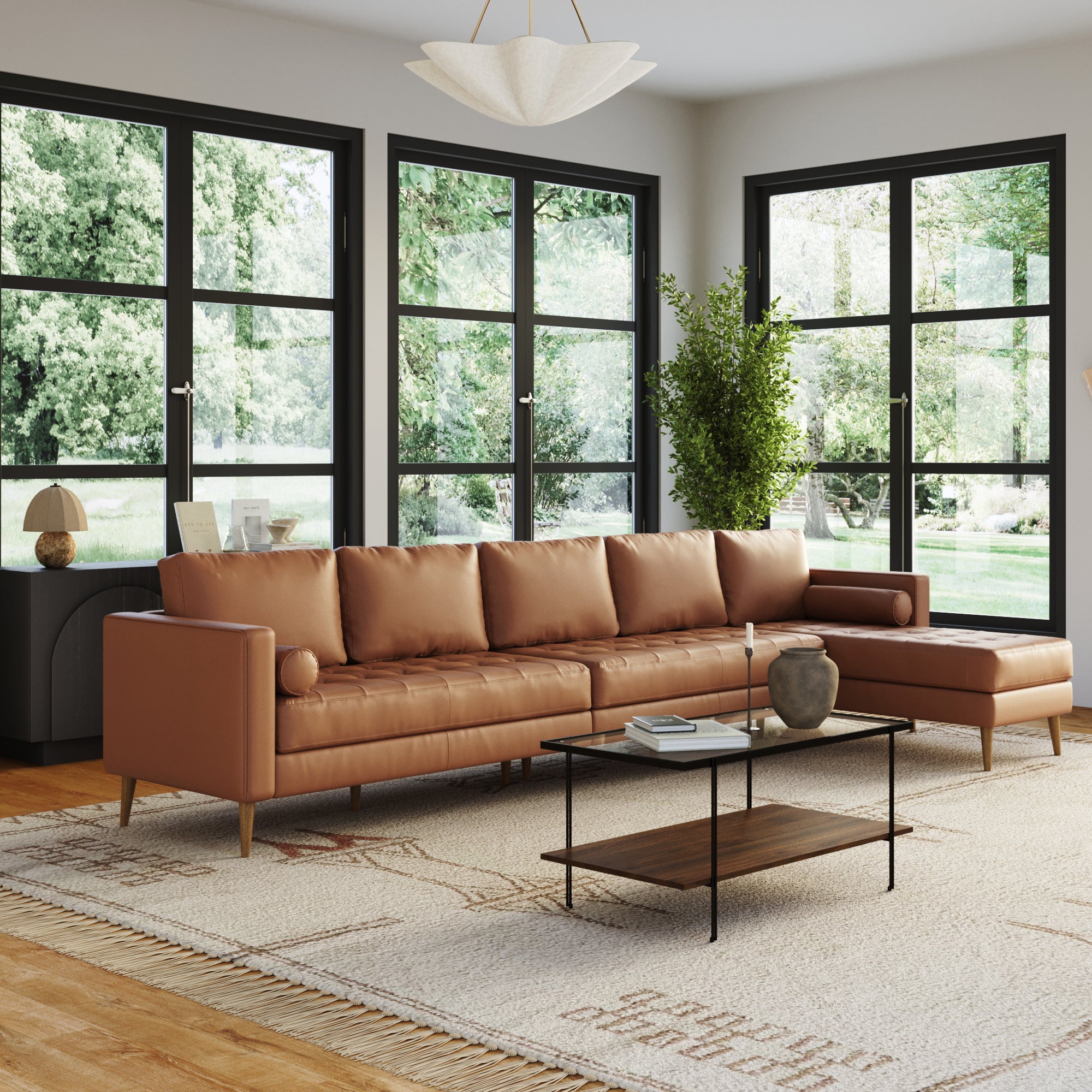 A modern living room with large windows, abundant natural light, lush greenery outside, and a Nathan James Mid-Century Tufted Leather 5-Seat Sofa with Bench Seat & Chaise in camel, plus a black-framed glass coffee table, patterned rug, and indoor plants.