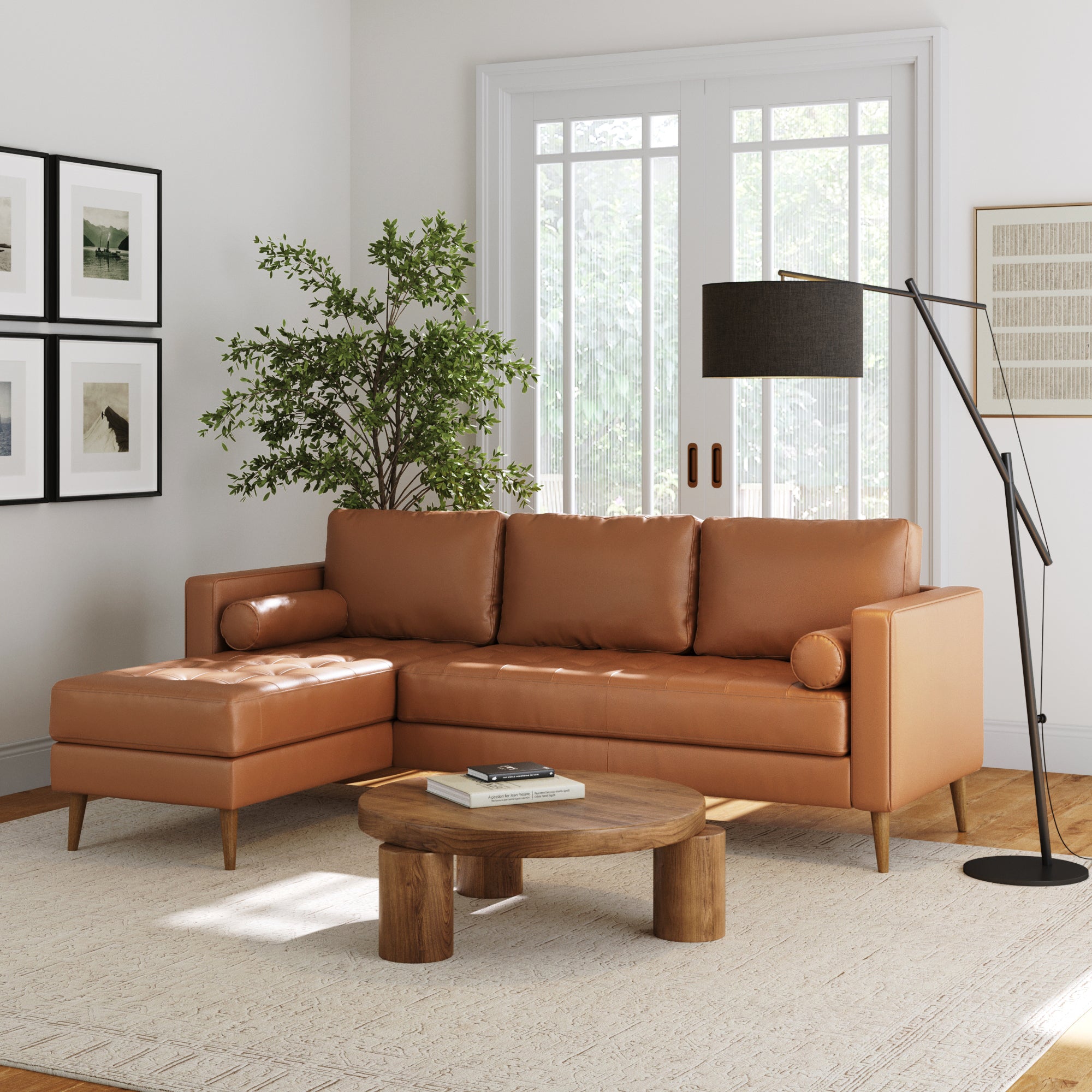 A modern living room with a Nathan James Mid-Century Tufted Leather Sofa Bench Seat in camel, round wooden coffee table, floor lamp, large potted plant, framed art, and sunlight streaming through white-curtained windows.