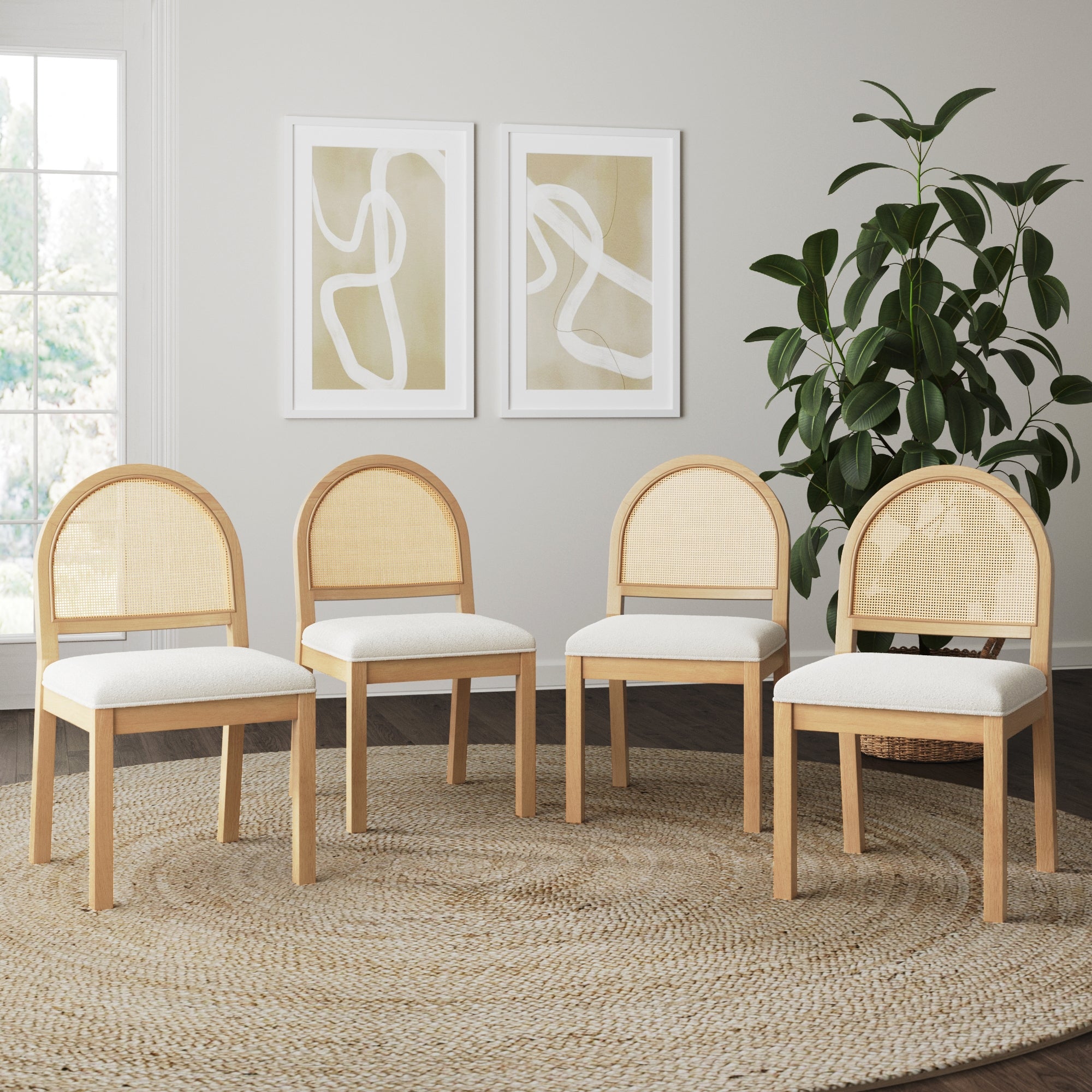Four Nathan James Boucle & Rattan Dining Chairs in Warm Pine (Set of 4) sit on a round woven cane rug in a bright room, with a large potted plant and two abstract framed artworks in the background.
