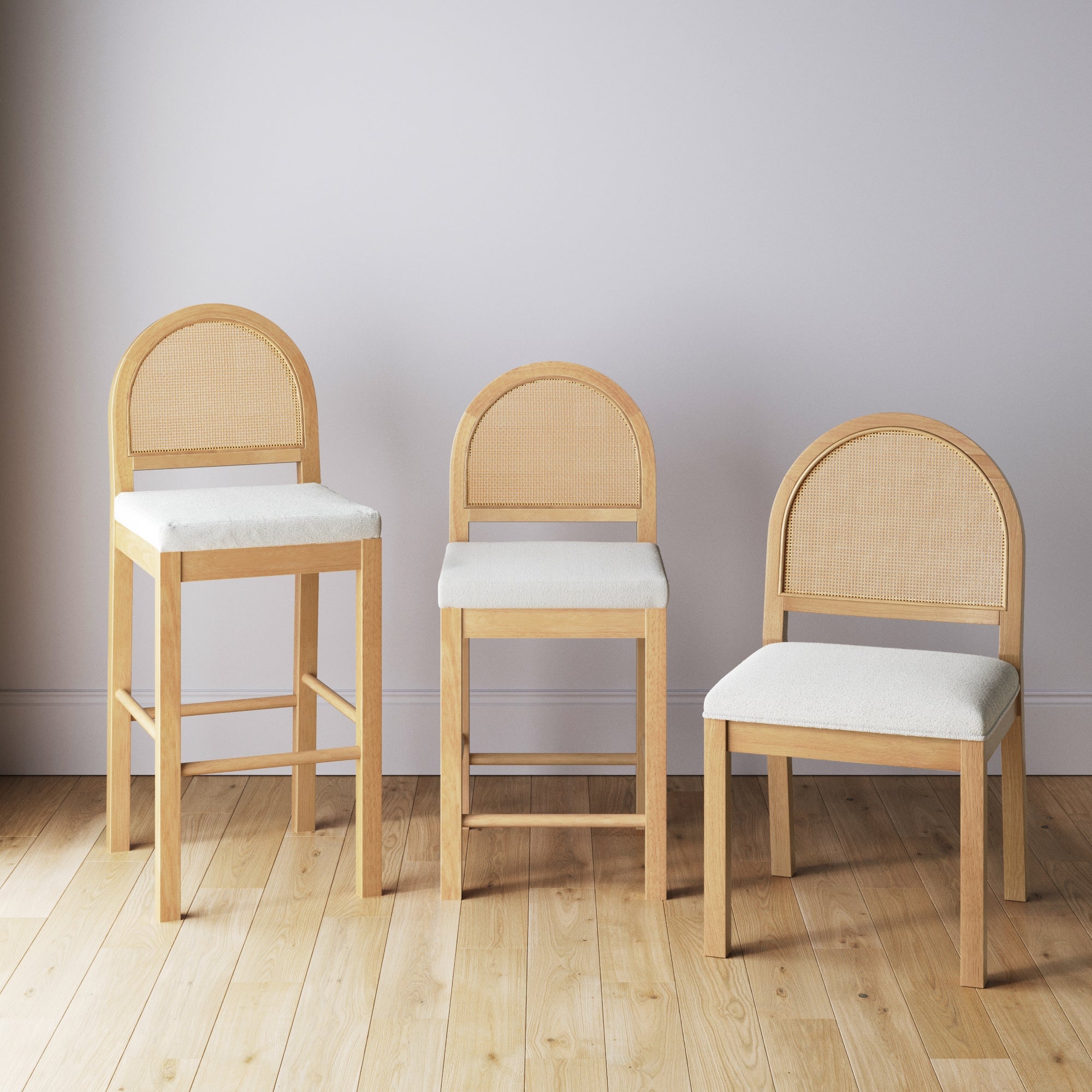 Three wooden chairs with rounded cane backs and light-colored cushioned seats, arranged side by side on a light wood floor against a plain, light gray wall. Each chair is a different height.