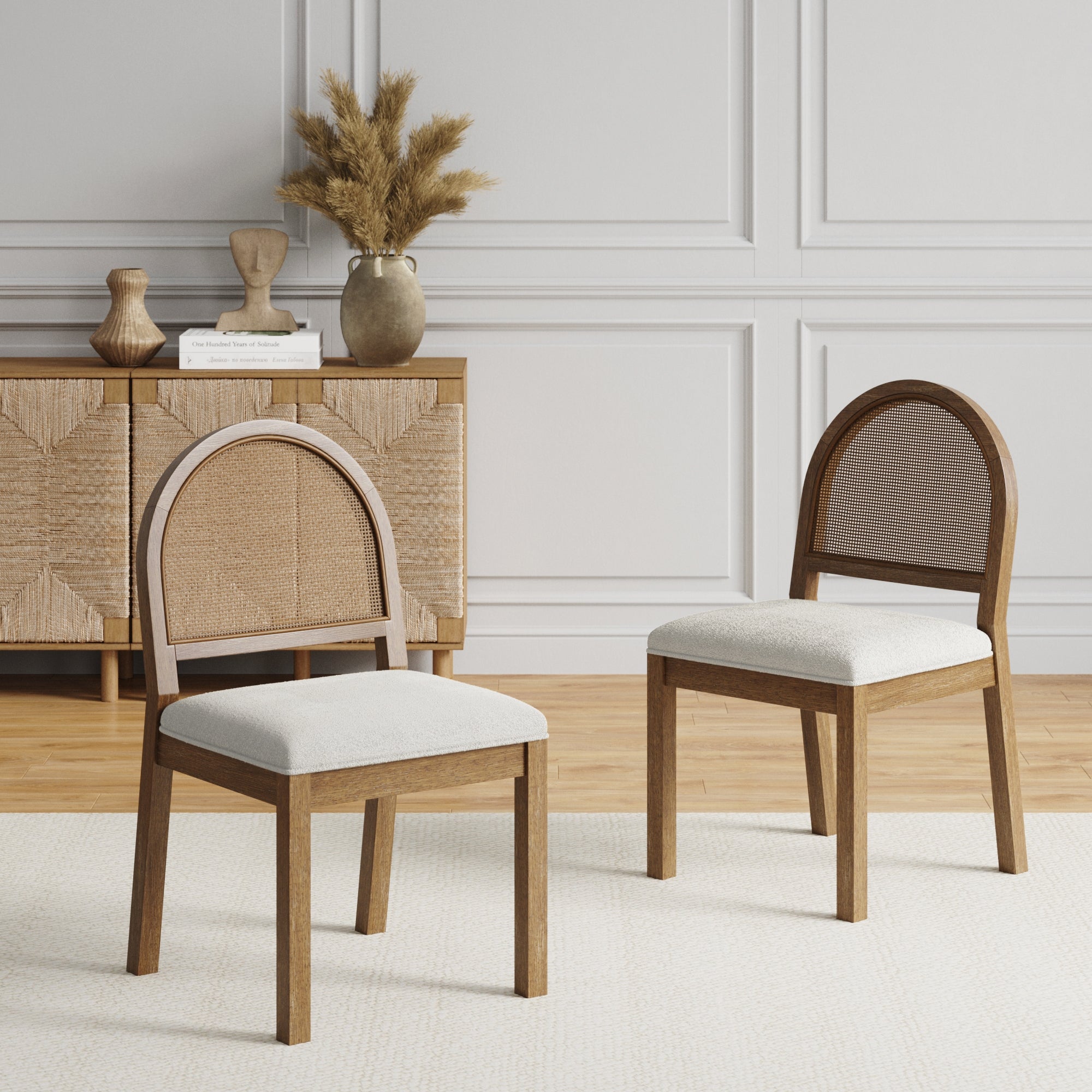 Two Nathan James Boucle & Rattan Dining Chairs in light brown (set of 2) with arched cane backs and upholstered seats sit on a rug in a modern room, with a cabinet, vases, and dried grasses against a paneled wall.