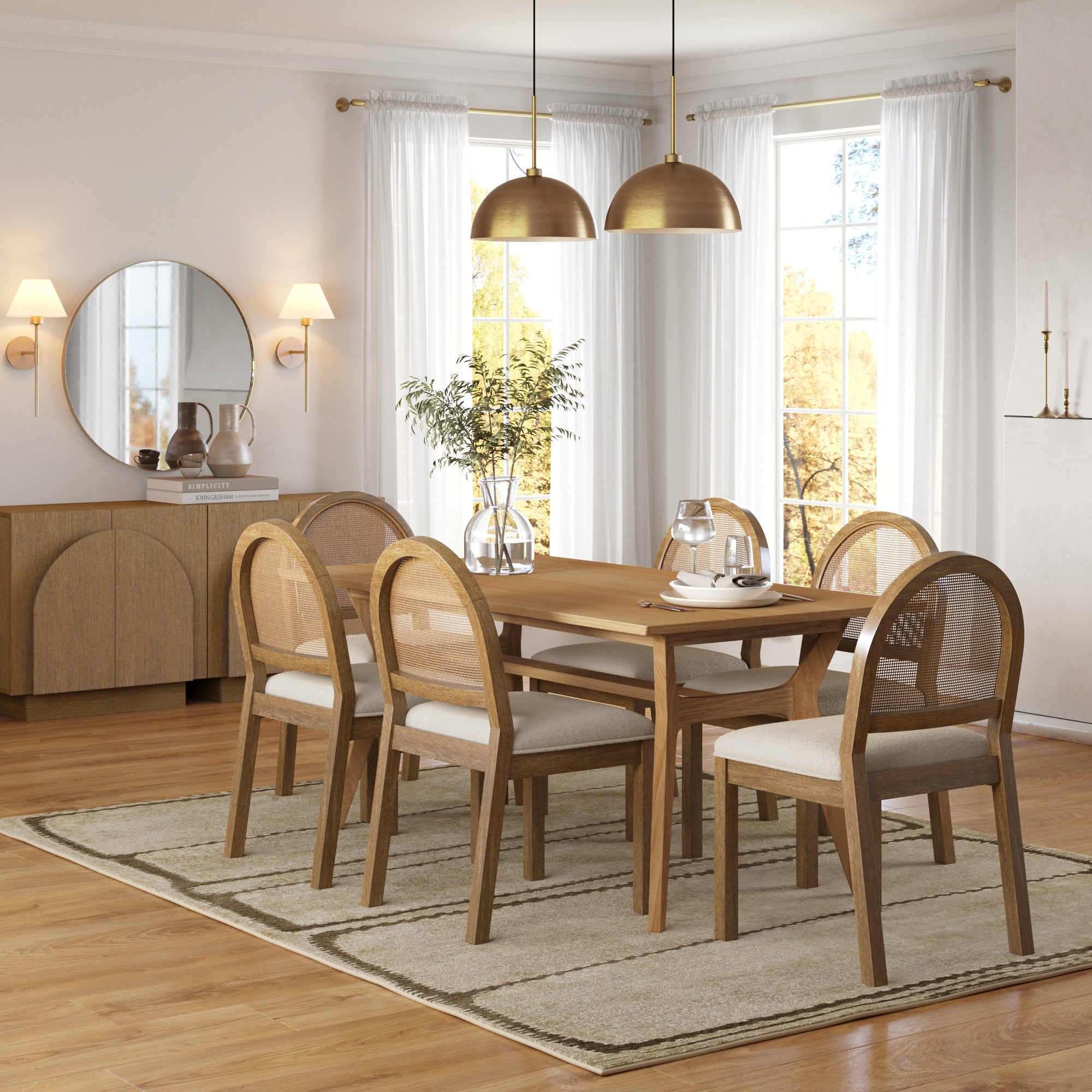 A modern dining room features a solid wood table with six Nathan James Boucle & Rattan Dining Chair Light Brown on a beige rug, two gold pendant lights overhead, a sideboard with a round mirror, and large windows with white curtains.