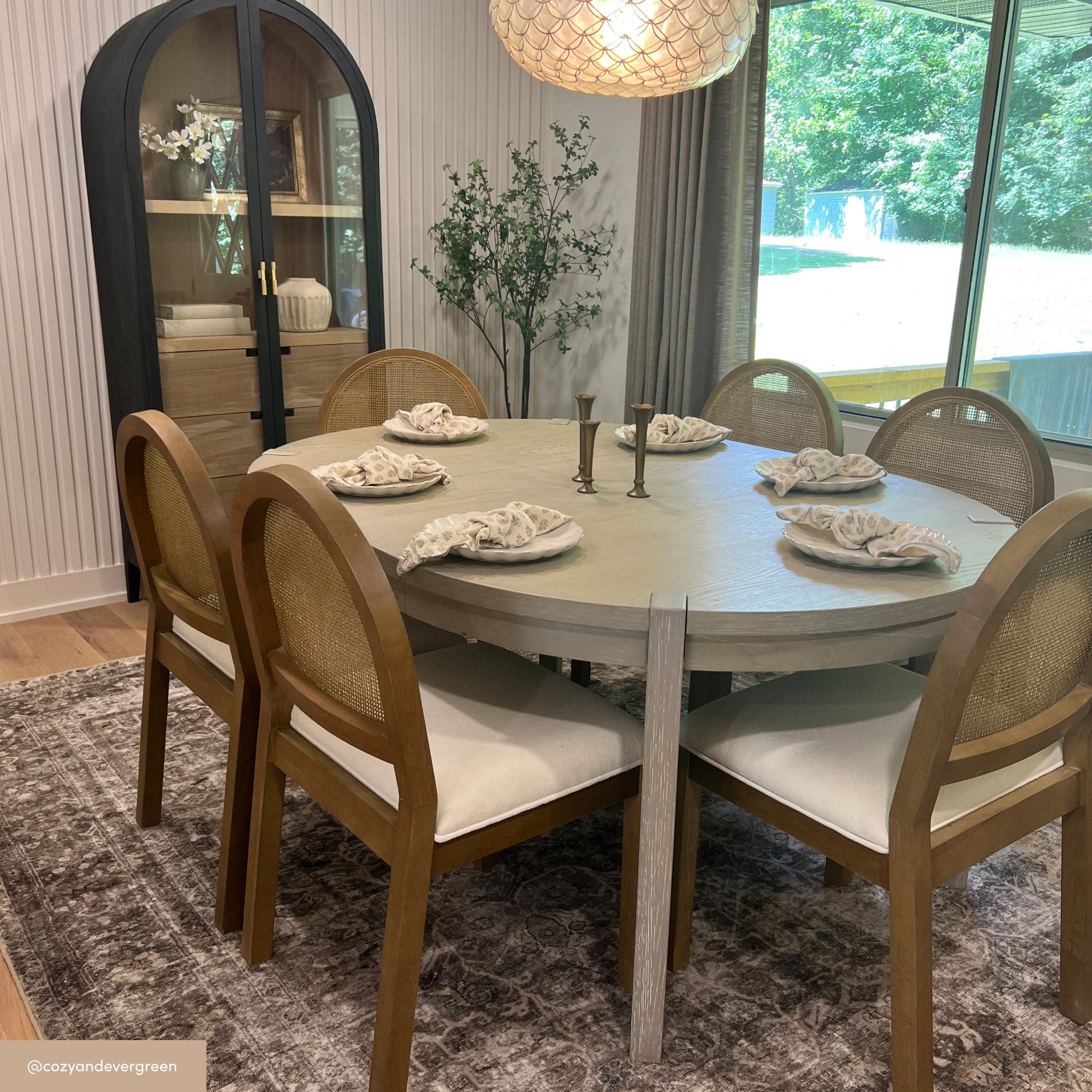 A round dining table with six wooden chairs is set with napkins and two candlesticks in a bright room, featuring a large window, patterned rug, black cabinet, and potted plant.