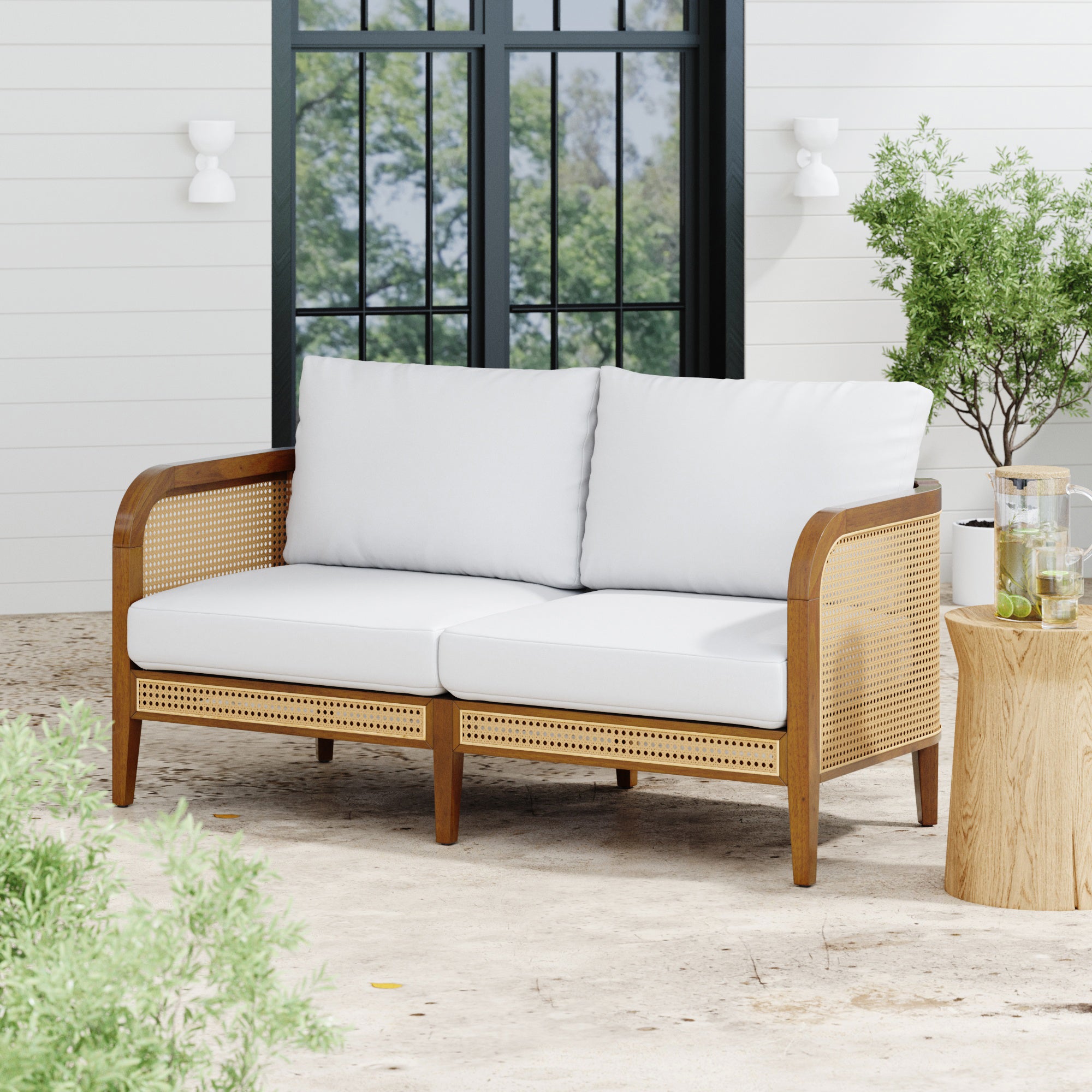 The Nathan James Outdoor Wood & Rattan Patio Loveseat Acacia, featuring removable cushion covers and a weatherproof acacia wood frame, sits on a patio near a wood side table with drinks, surrounded by greenery in front of a white paneled wall and window.