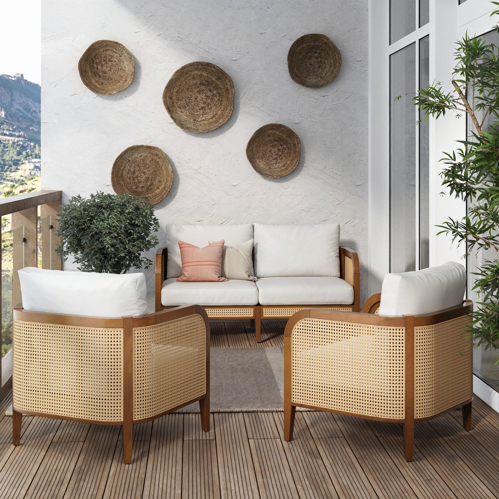 A modern balcony features the Nathan James Outdoor 3 Piece Wood Patio Set with rattan loveseat and chairs, white cushions, a potted plant, rug, woven baskets as wall decor, and mountain views from the wooden railing.