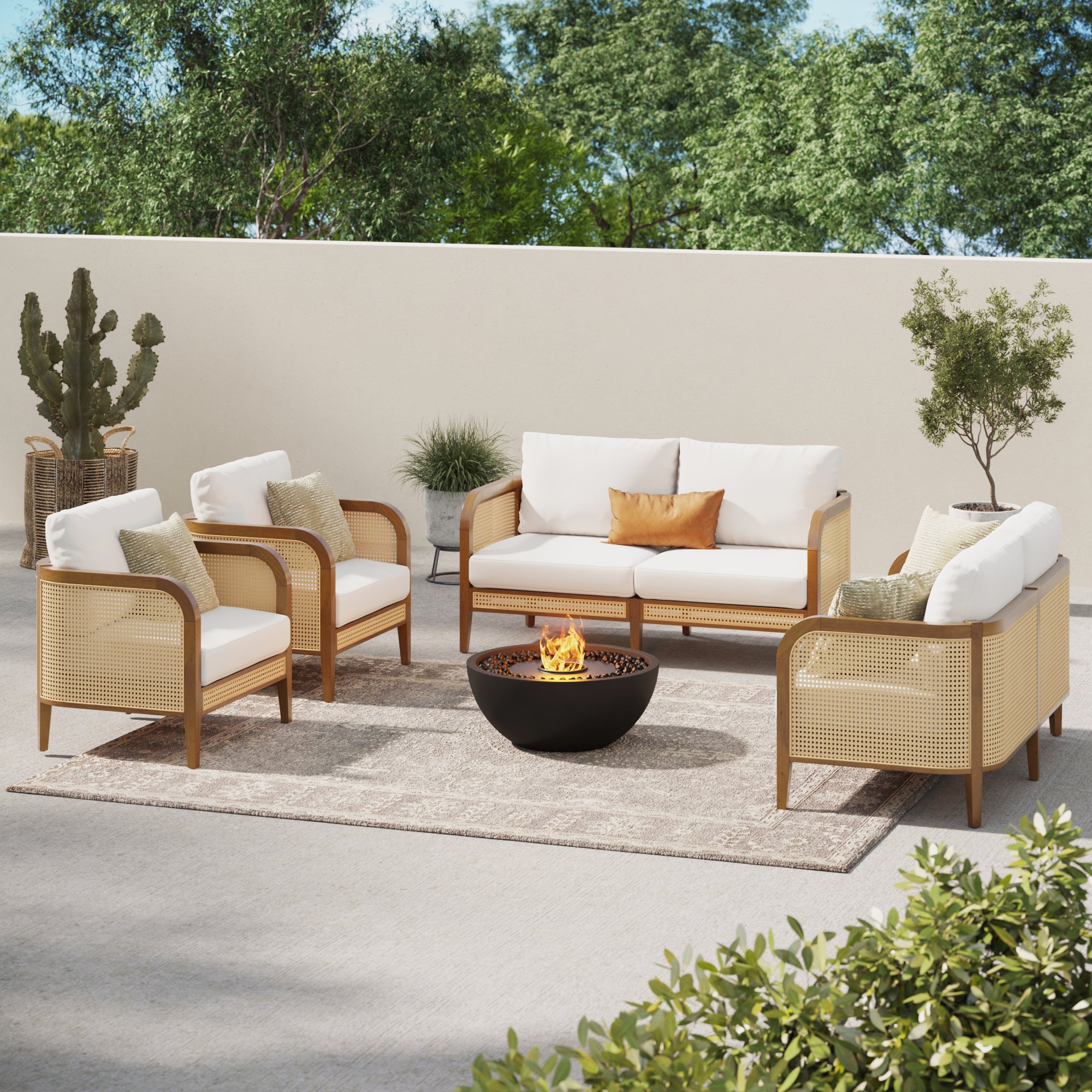 A modern patio with the Nathan James Outdoor 4 Piece Wood Patio Set—rattan loveseats and chairs—on a rug with a round fire pit, potted plants, white wall, and green trees under a clear sky.