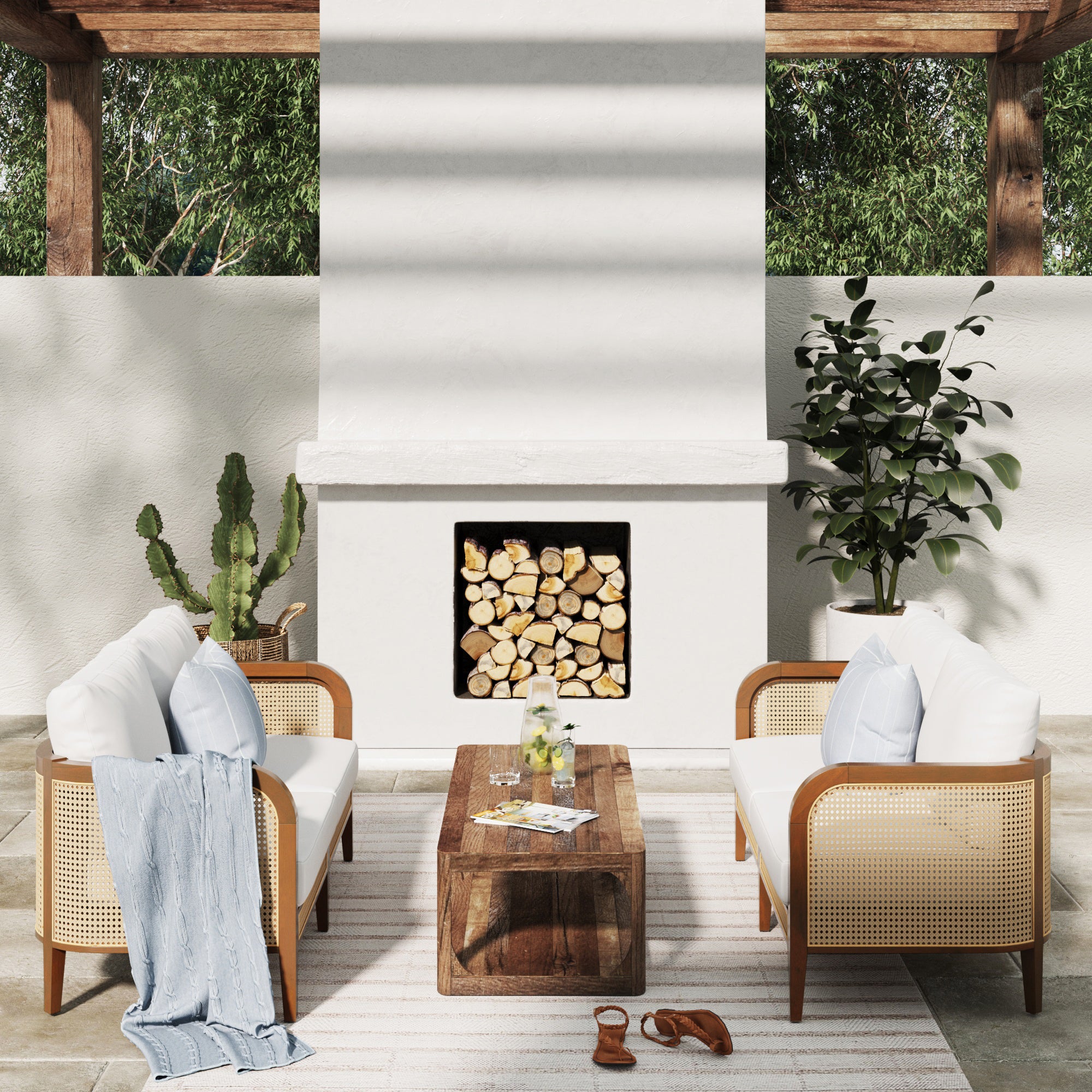 Outdoor seating area with two cushioned armchairs, a wooden coffee table, and a fireplace with stacked firewood. Plants are beside the fireplace, and sunlight casts shadows across the space.