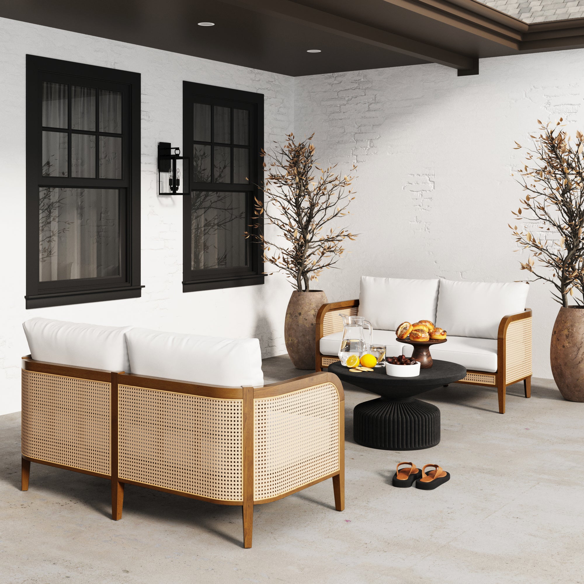 A modern patio with two rattan sofas with white cushions, a round black table with breakfast items, two large potted trees, and two pairs of sandals on a light concrete floor against a white brick wall.