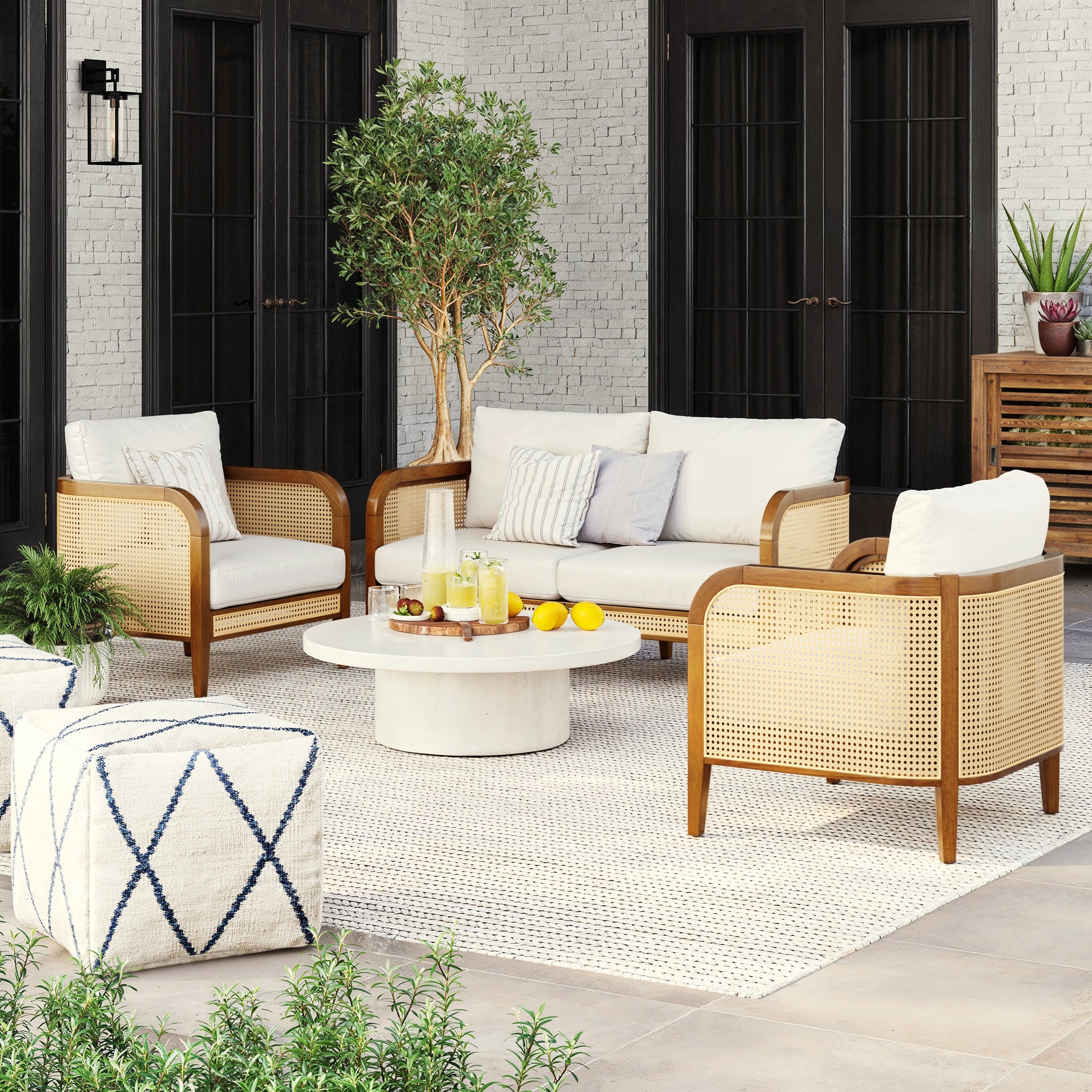 A modern patio features the Nathan James Outdoor 3 Piece Wood Patio Set with a rattan loveseat and chairs, cream cushions, a round coffee table for drinks, a white rug, potted plants, and black French doors against a light brick wall.