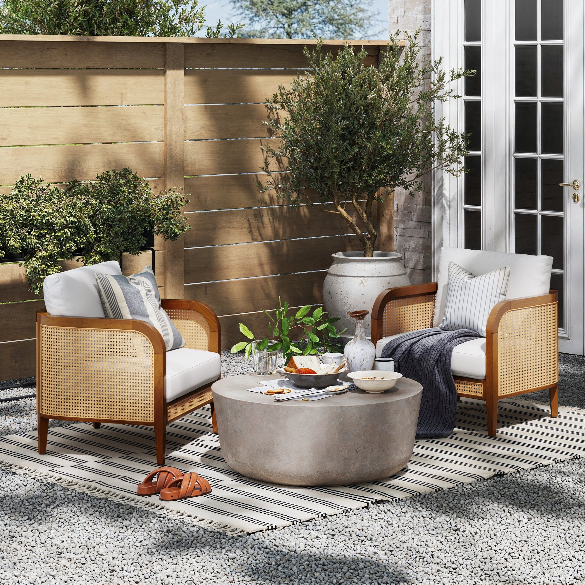 Two Nathan James Outdoor Wood & Rattan Patio Arm Chairs (Set of 2) with throw pillows face a round stone coffee table with drinks and snacks, all arranged on a striped rug in a gravel patio accented by potted plants and wooden fencing.