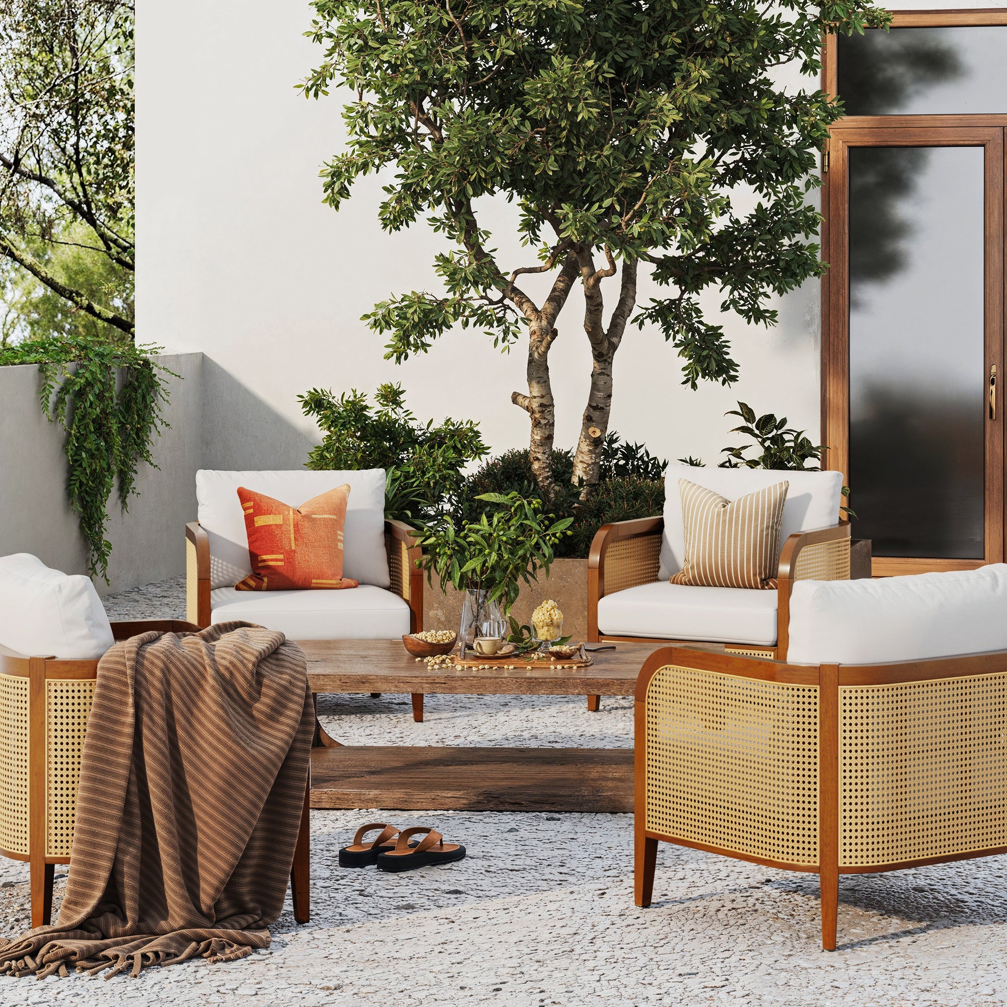A cozy outdoor patio with four Nathan James Outdoor Wood & Rattan Patio Arm Chairs Acacia (Set of 4) featuring cushions and pillows, a low wooden table with drinks, a potted tree, striped blanket, and sandals by a glass door on stone flooring.