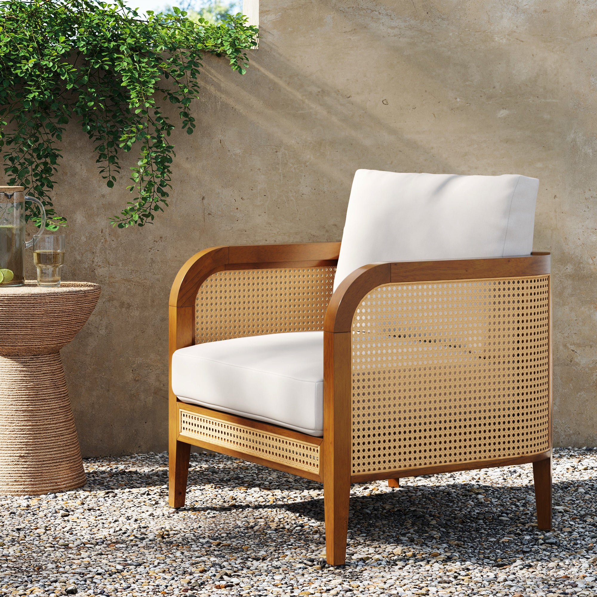 The Nathan James Outdoor Wood & Rattan Patio Arm Chair Acacia features a cane back and sides, white cushions, and weatherproof acacia wood. It sits beside drinks on a round woven table near green plants against a tan wall.