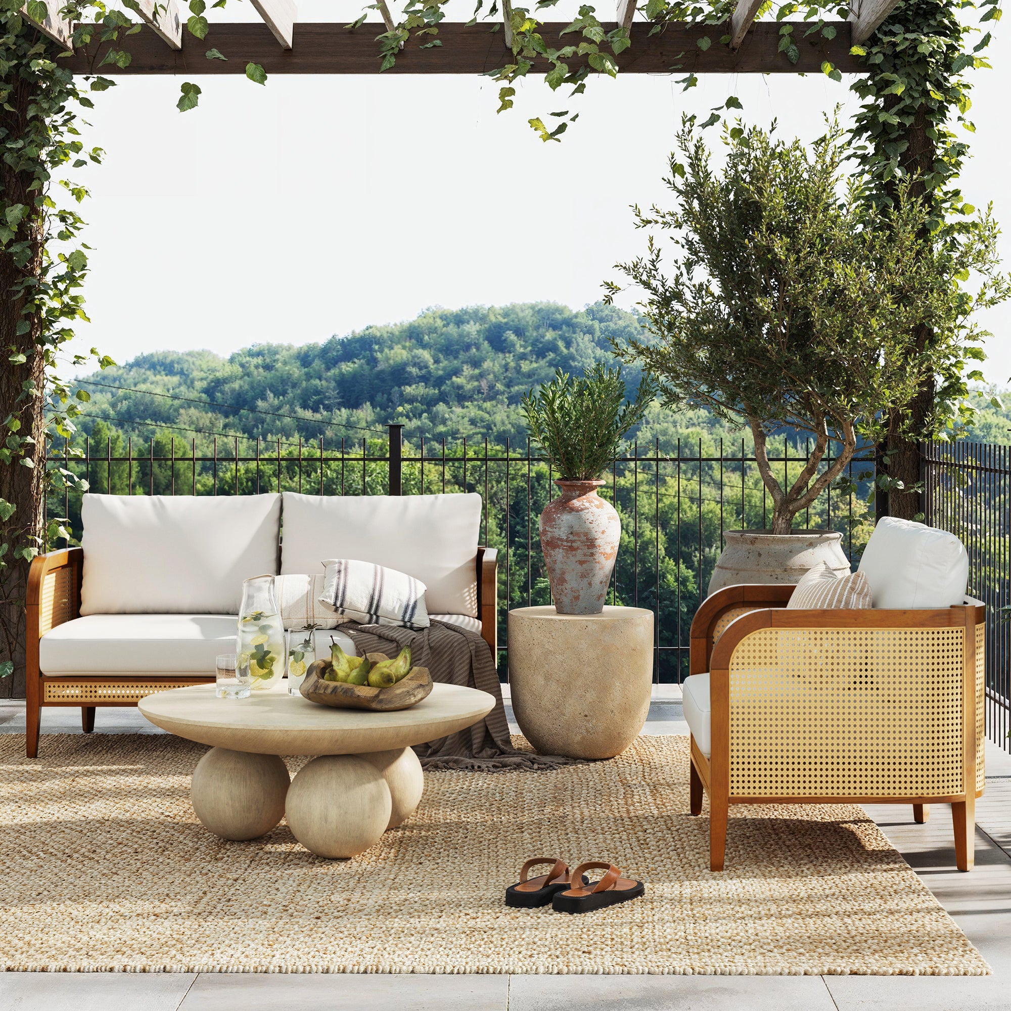Nathan James Outdoor Wood & Rattan Patio Arm Chair Acacia sits on a woven rug with white cushions, beside two rattan sofas, a round coffee table with fruit and drinks, potted plants, and sandals under a pergola overlooking lush hills.