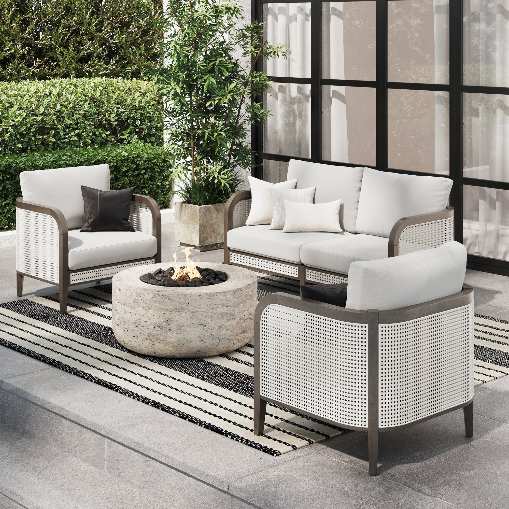 Modern outdoor patio set with two armchairs, a loveseat, and a round stone fire pit on a striped rug, surrounded by greenery and next to tall windows. Cushions are white with neutral-toned throw pillows.