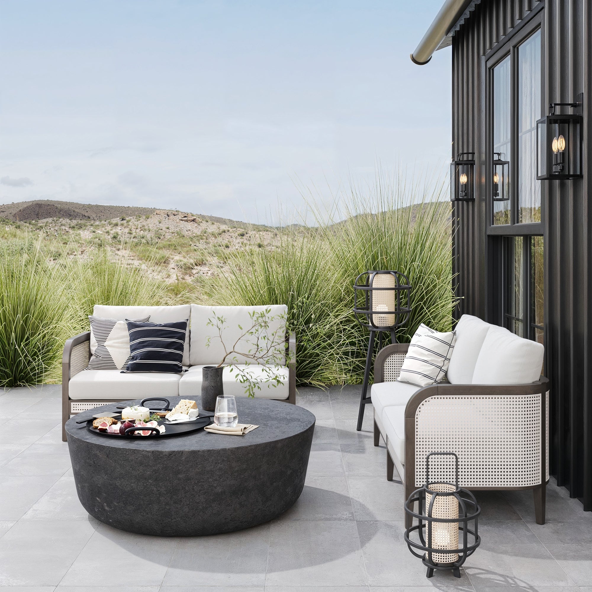 Modern patio with two Nathan James Outdoor Wood & Rattan Patio Loveseats in Graphite, washable cushions, a round black coffee table with snacks, potted plants, and lanterns—set against a black house framed by tall grasses and hills.