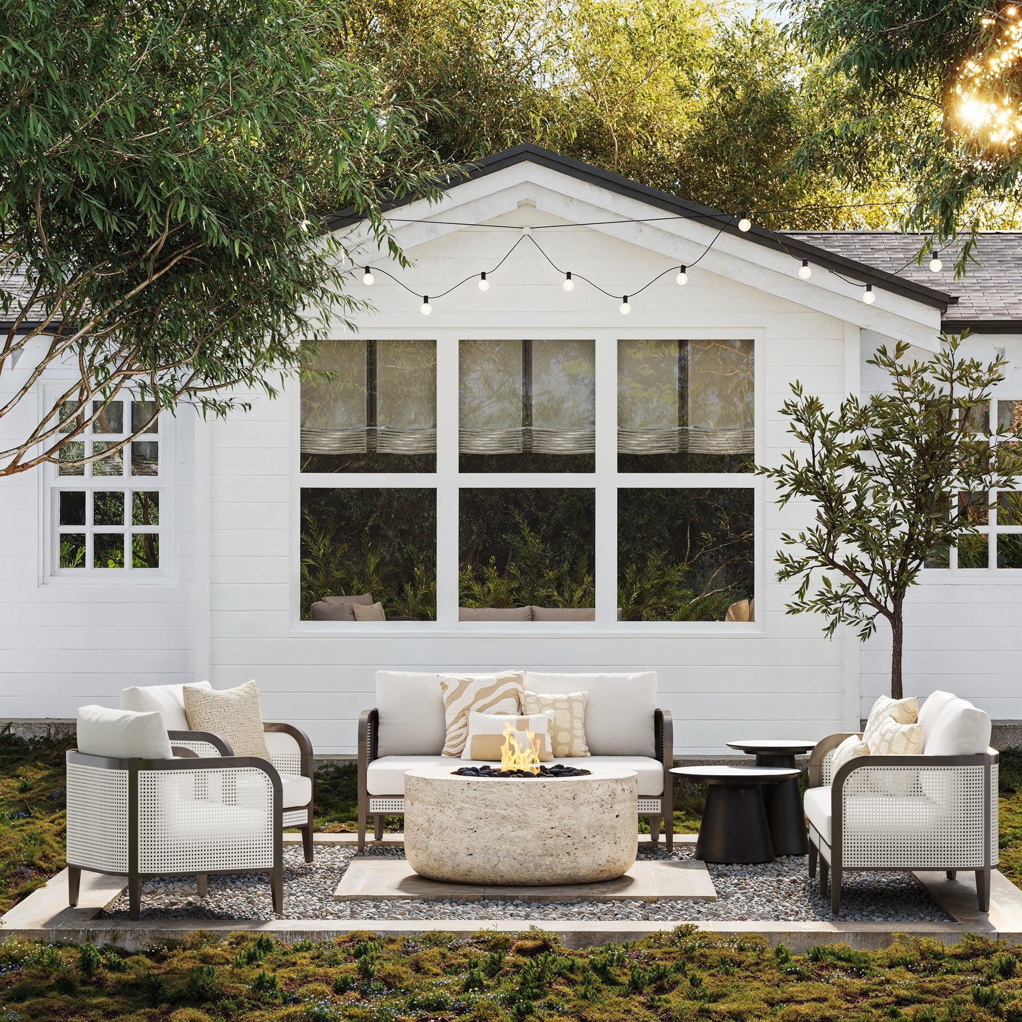 A cozy patio showcases the Nathan James Outdoor 4 Piece Wood Patio Set with rattan loveseats and chairs arranged around a stone fire pit, under string lights, beside a white house with large windows and lush greenery.