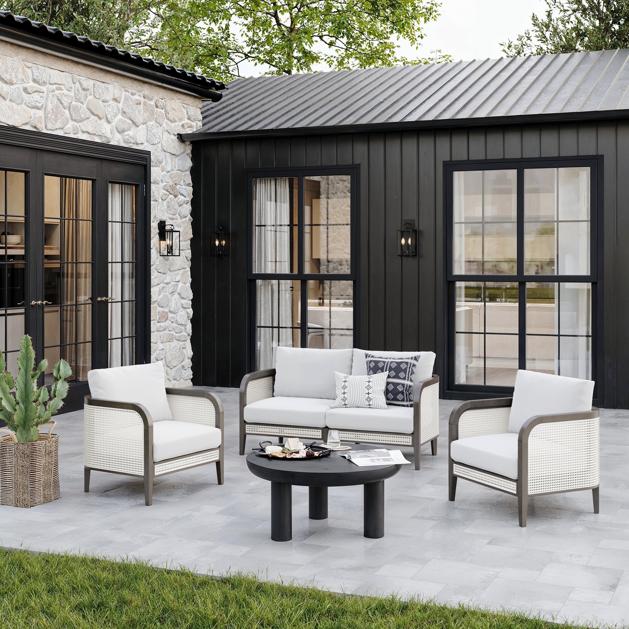 The patio features the Nathan James Outdoor Wood & Rattan Patio Loveseat in graphite, paired with two armchairs and a round black coffee table. White washable cushions and a basket cactus accent the space, framed by dark siding and large windows.