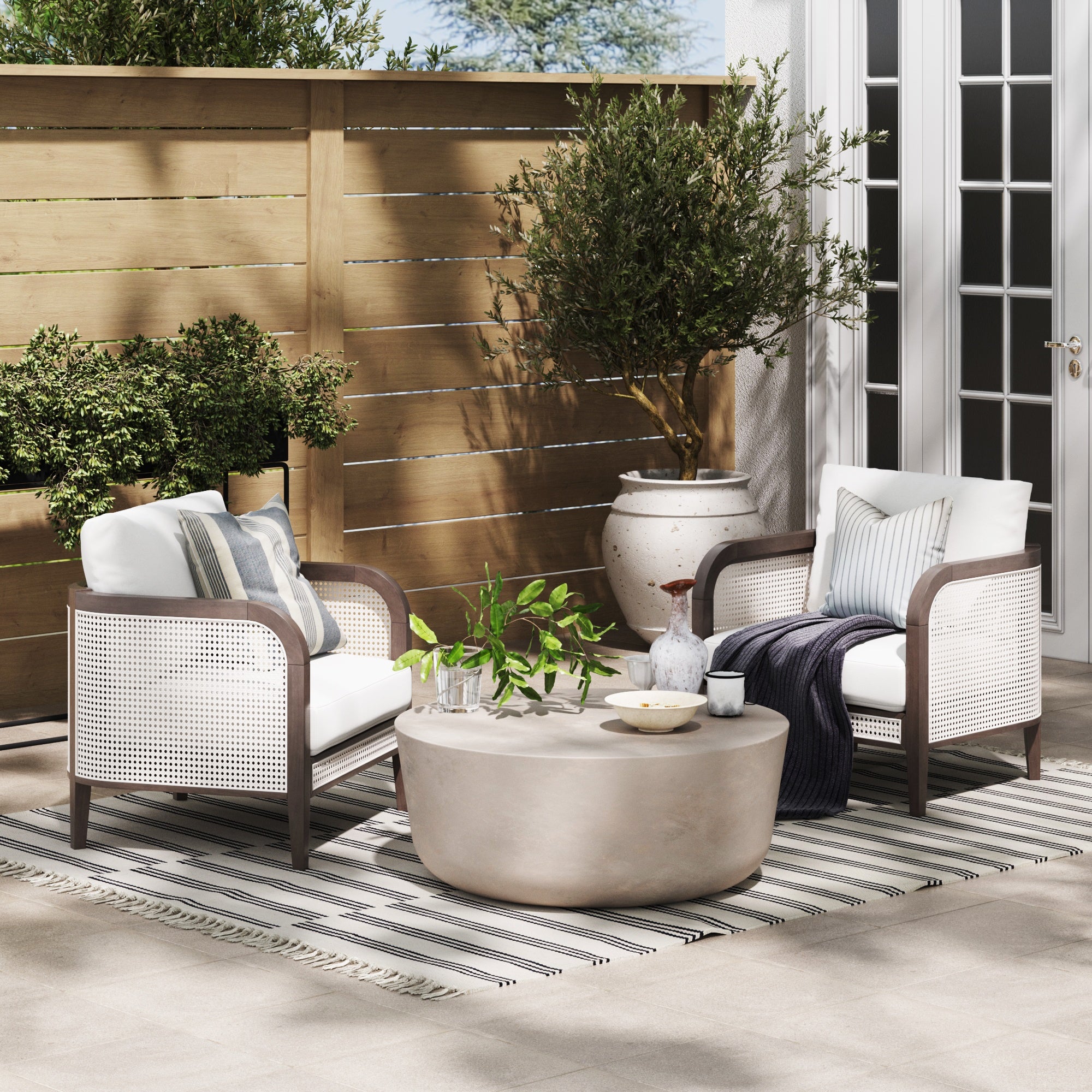 A cozy patio setting with two cushioned armchairs, a round coffee table decorated with plants, and a striped rug. Wooden fencing, potted plants, and French doors complete the relaxing outdoor space.