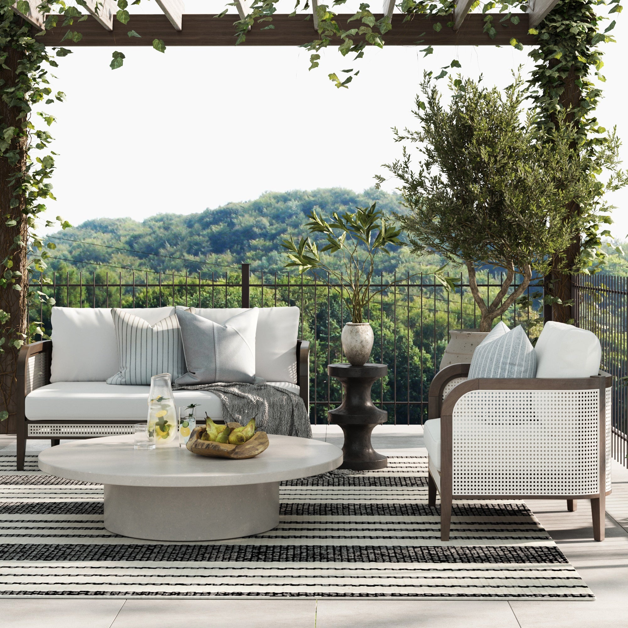 A stylish patio features Nathan James Outdoor Wood & Rattan Patio Arm Chair in Graphite with washable cushion covers, a round coffee table with drinks and fruit, lush potted plants, and a vine-draped pergola overlooking green hills.