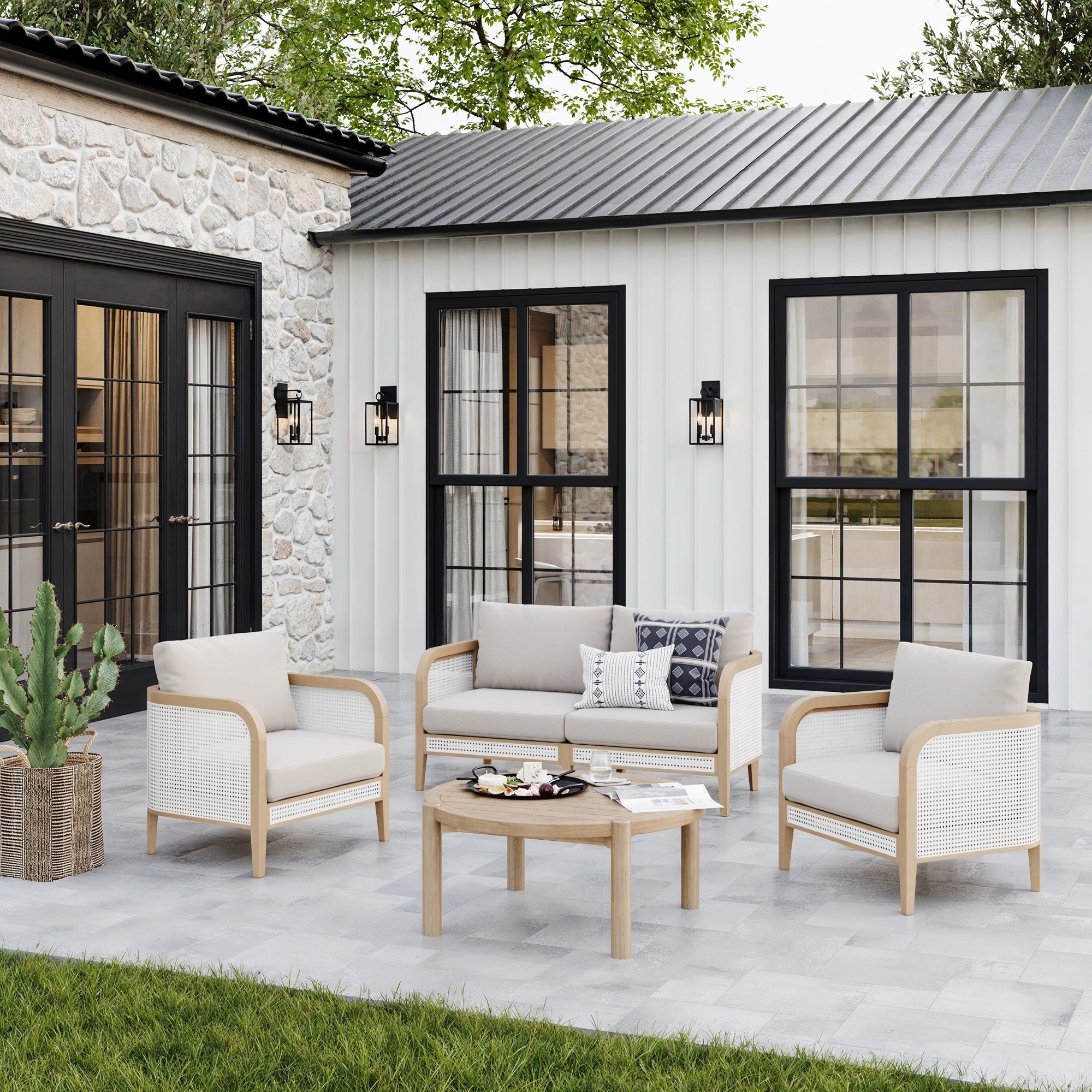 A modern patio showcases the Nathan James Outdoor 3 Piece Wood Patio Set Rattan Loveseat and Chairs with white cushions, set against a stone and white-paneled house featuring striking black-trimmed windows.