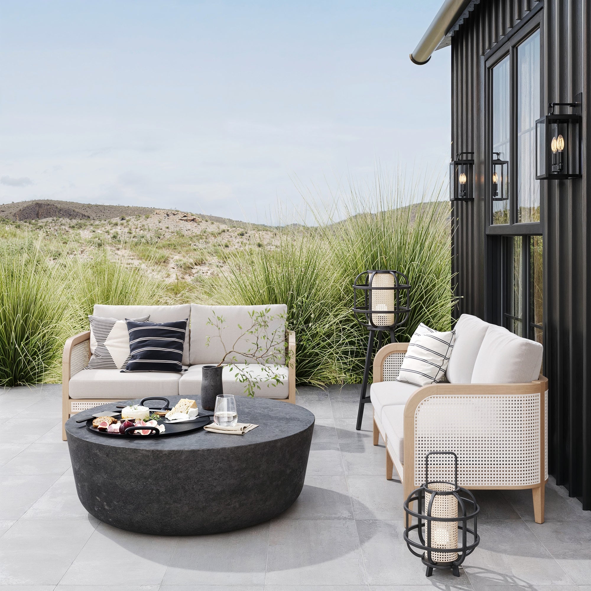 Outdoor patio with two Nathan James Outdoor Wood & Rattan Patio Loveseats in Gray Oak, washable cushions, a large round black table with drinks and snacks, floor lanterns, and a desert landscape with hills and grass beyond.