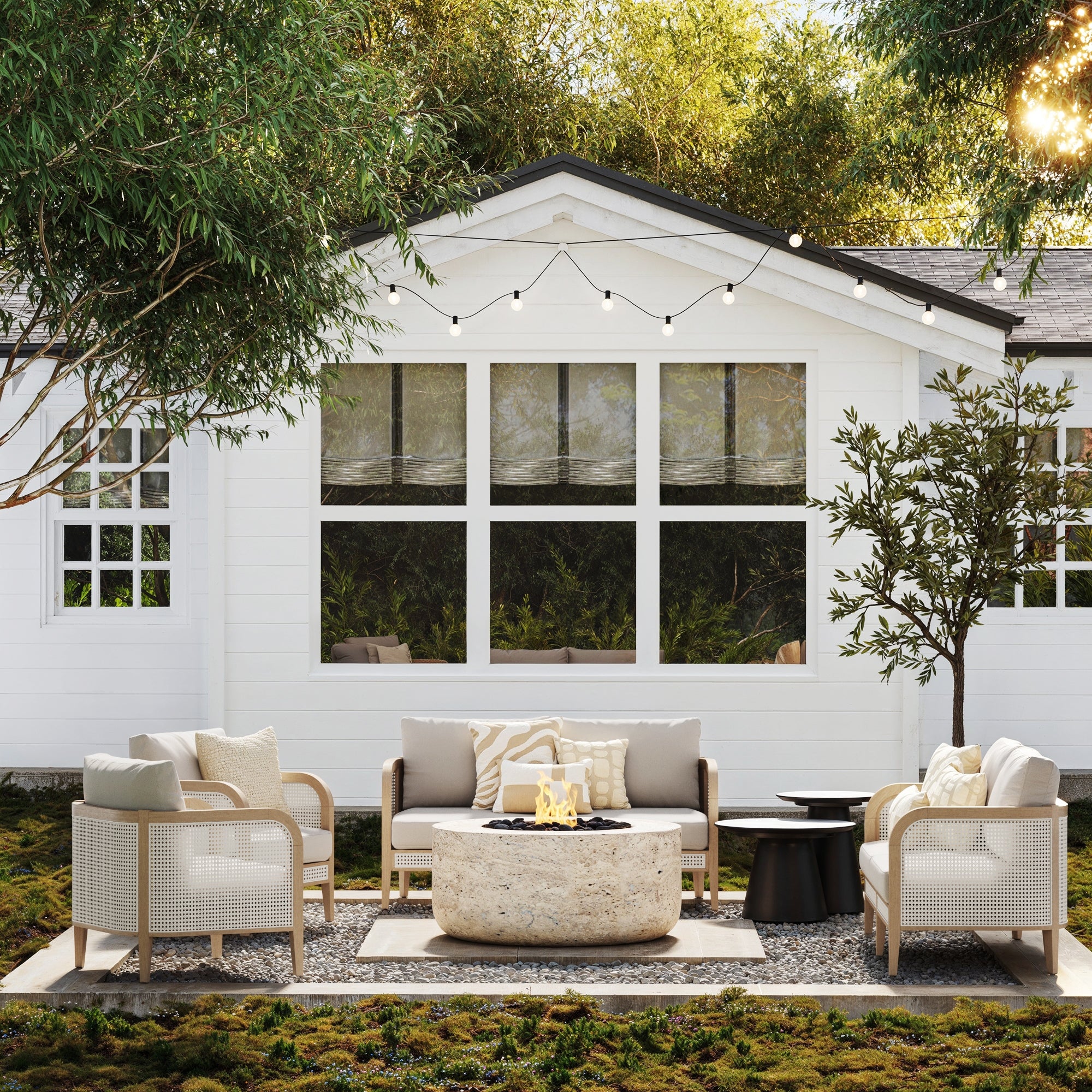 A contemporary patio features the Nathan James Outdoor 4 Piece Wood Patio Set with rattan loveseats and chairs, a round stone fire pit, string lights above, and abundant greenery around a white house with large windows.