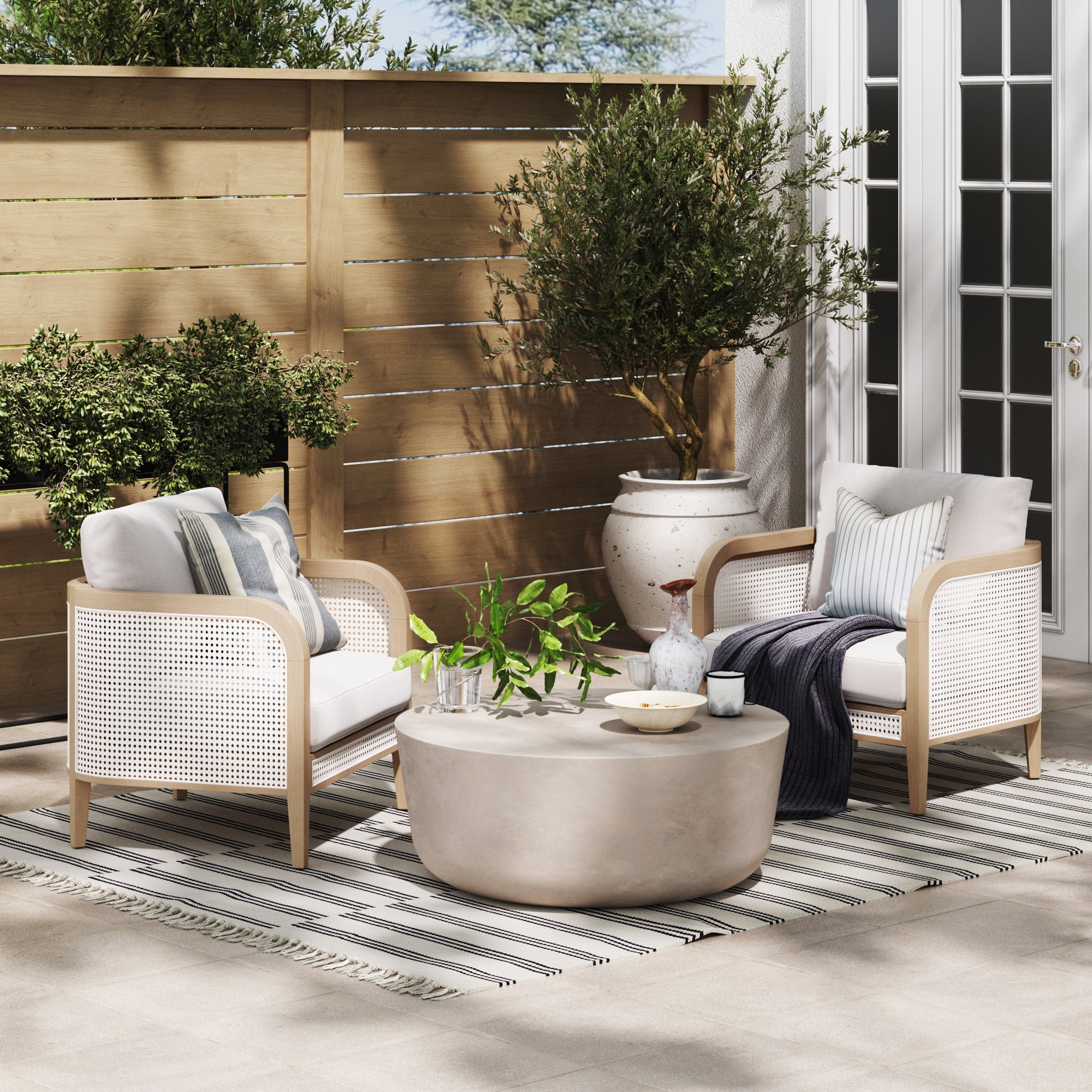 Two cushioned armchairs with striped pillows sit by a round coffee table topped with plants and decor, on a striped rug in an outdoor patio area with wooden fencing, potted trees, and glass doors.