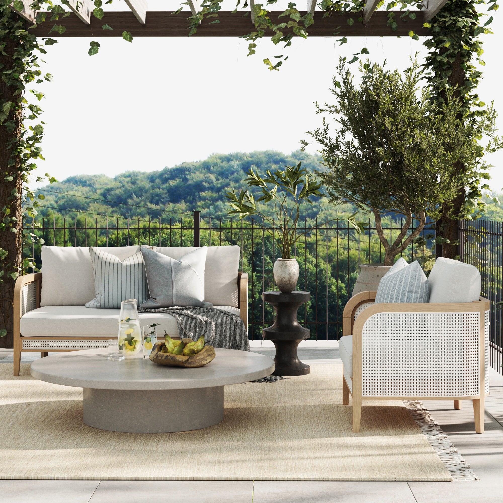 Outdoor patio with two cushioned chairs, a round coffee table with a water pitcher and fruit, a small side table, potted plants, a rug, and a scenic view of green hills in the background.