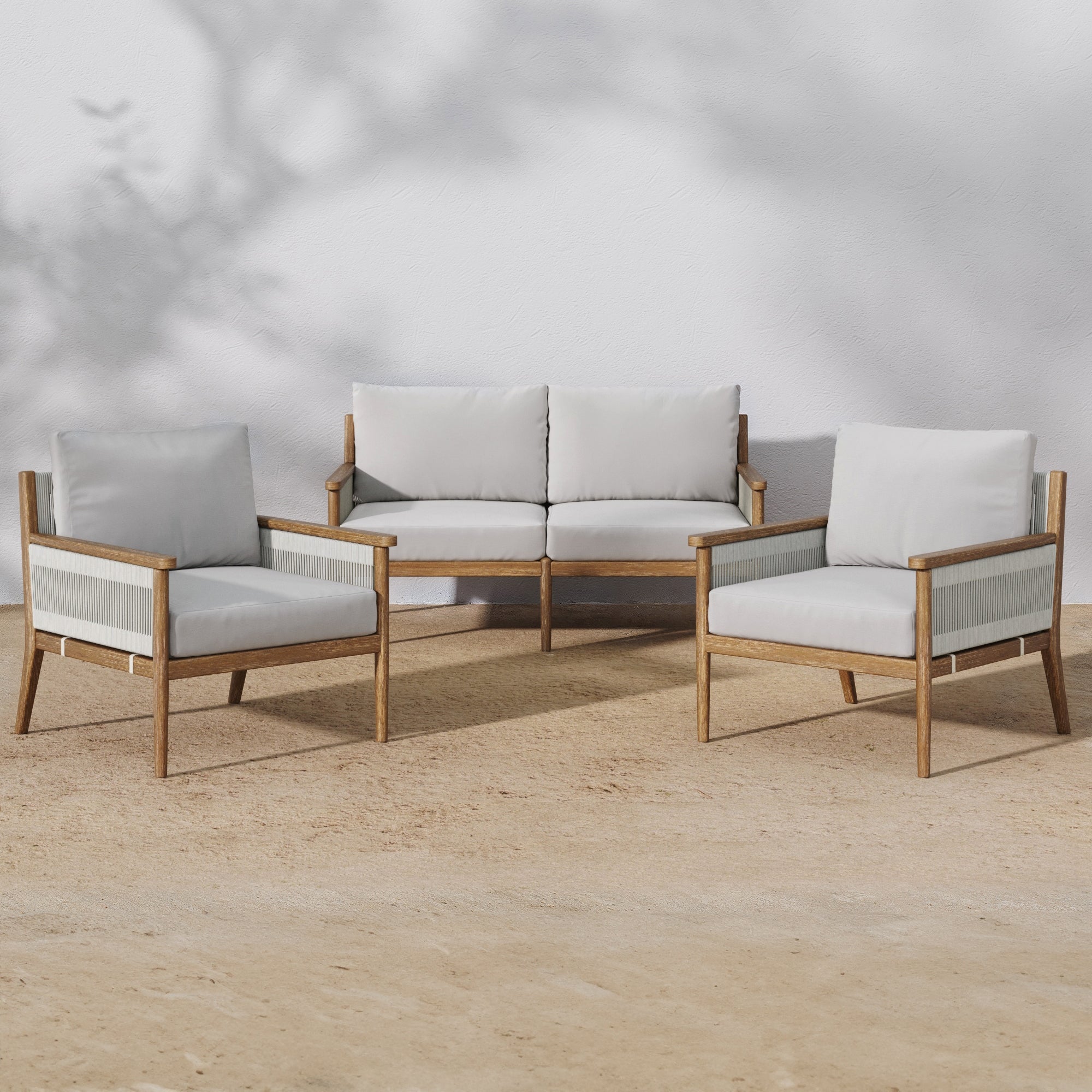 The Nathan James Outdoor 3 Piece Wood Patio Set, featuring a loveseat and chairs with weatherproof acacia wood frames and soft white washable cushions, is arranged on sand against a white wall with shadowy tree branches.