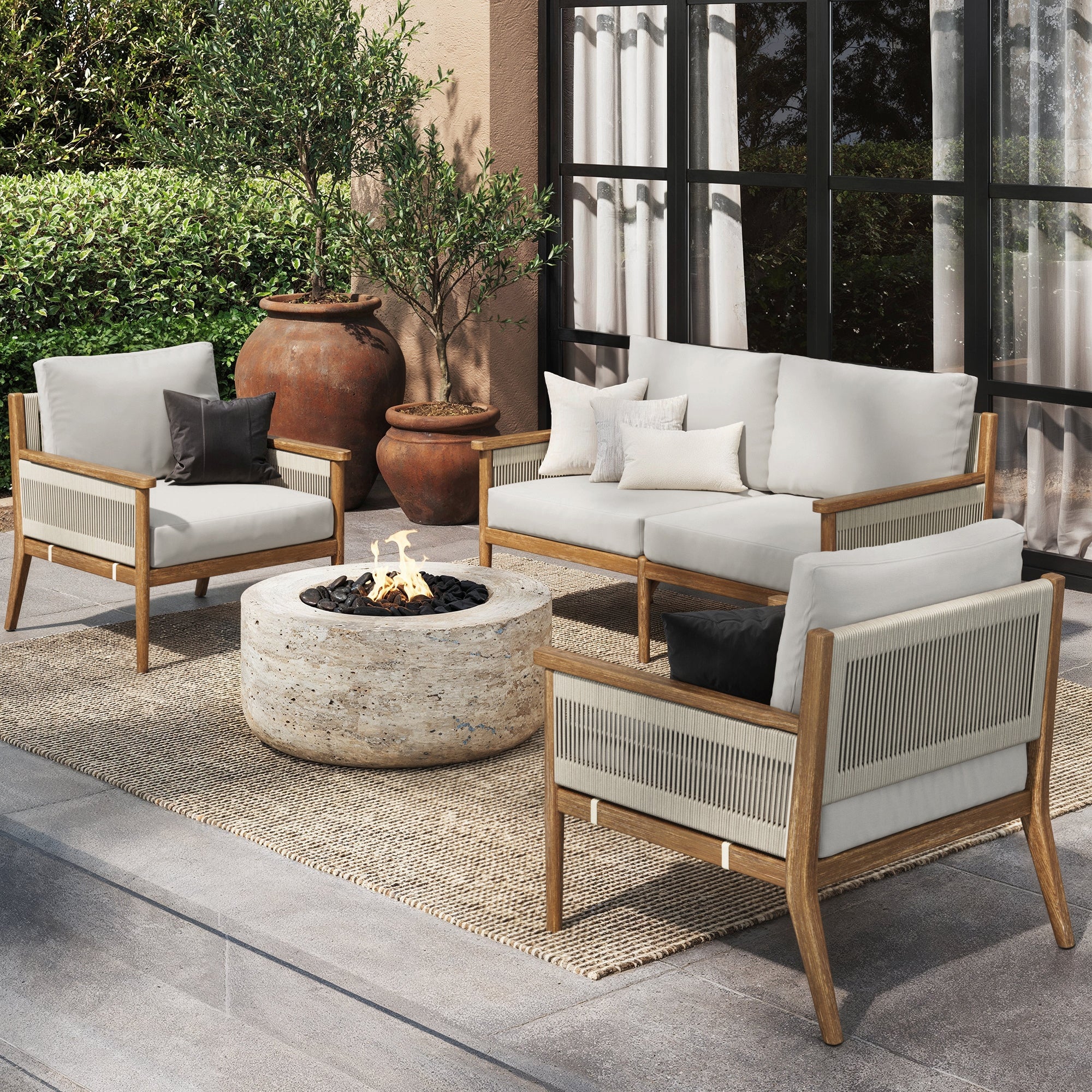 A modern patio showcases the Nathan James Outdoor 3 Piece Wood Patio Set Loveseat and Chairs, with weatherproof frames, washable light cushions, black and white accent pillows, a stone fire pit, terra cotta planters, greenery, and sleek glass doors.