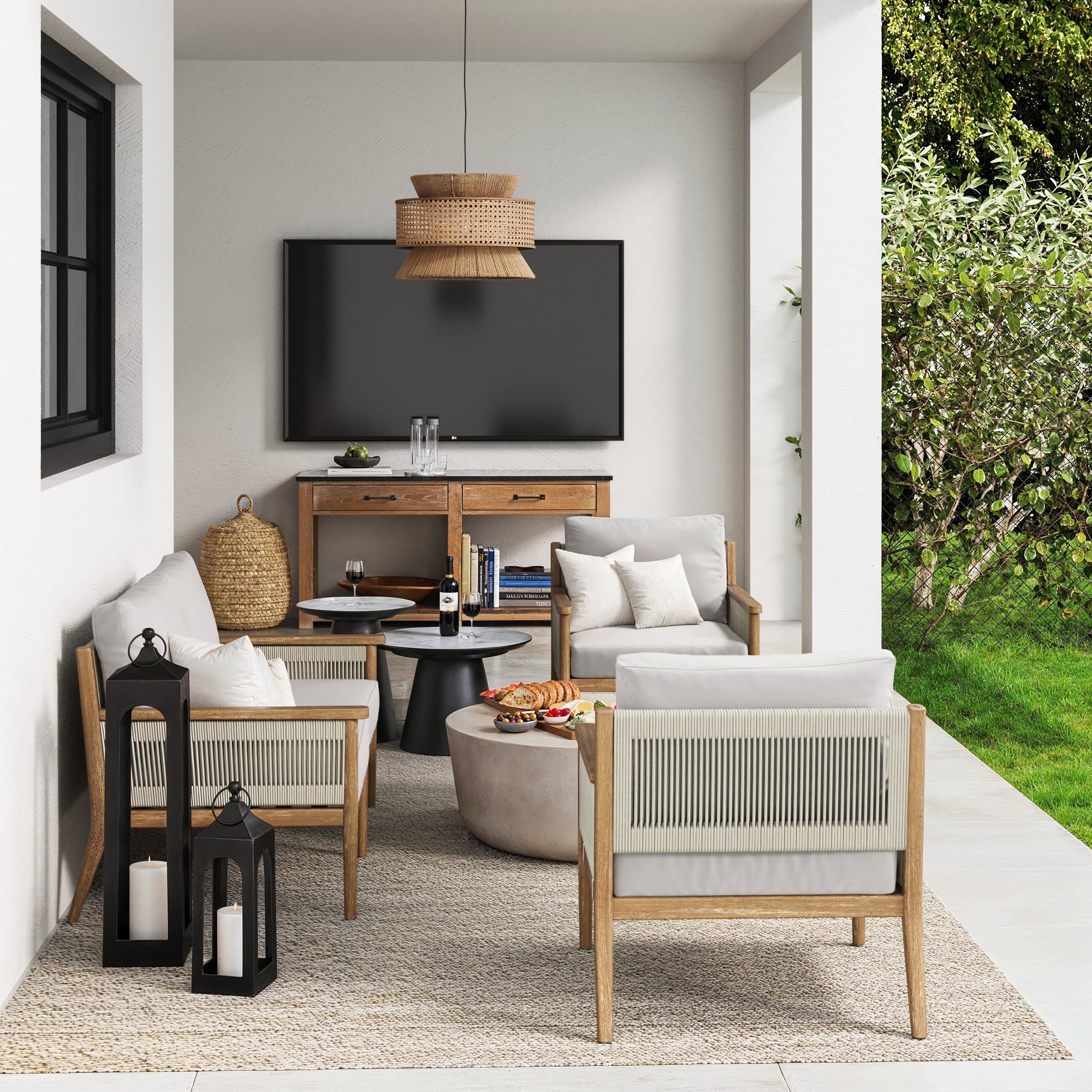 A cozy covered patio with cushioned wooden chairs, a sofa, a round ottoman, black lanterns, a TV on the wall, a wicker pendant light, and a view of a green yard with trees.