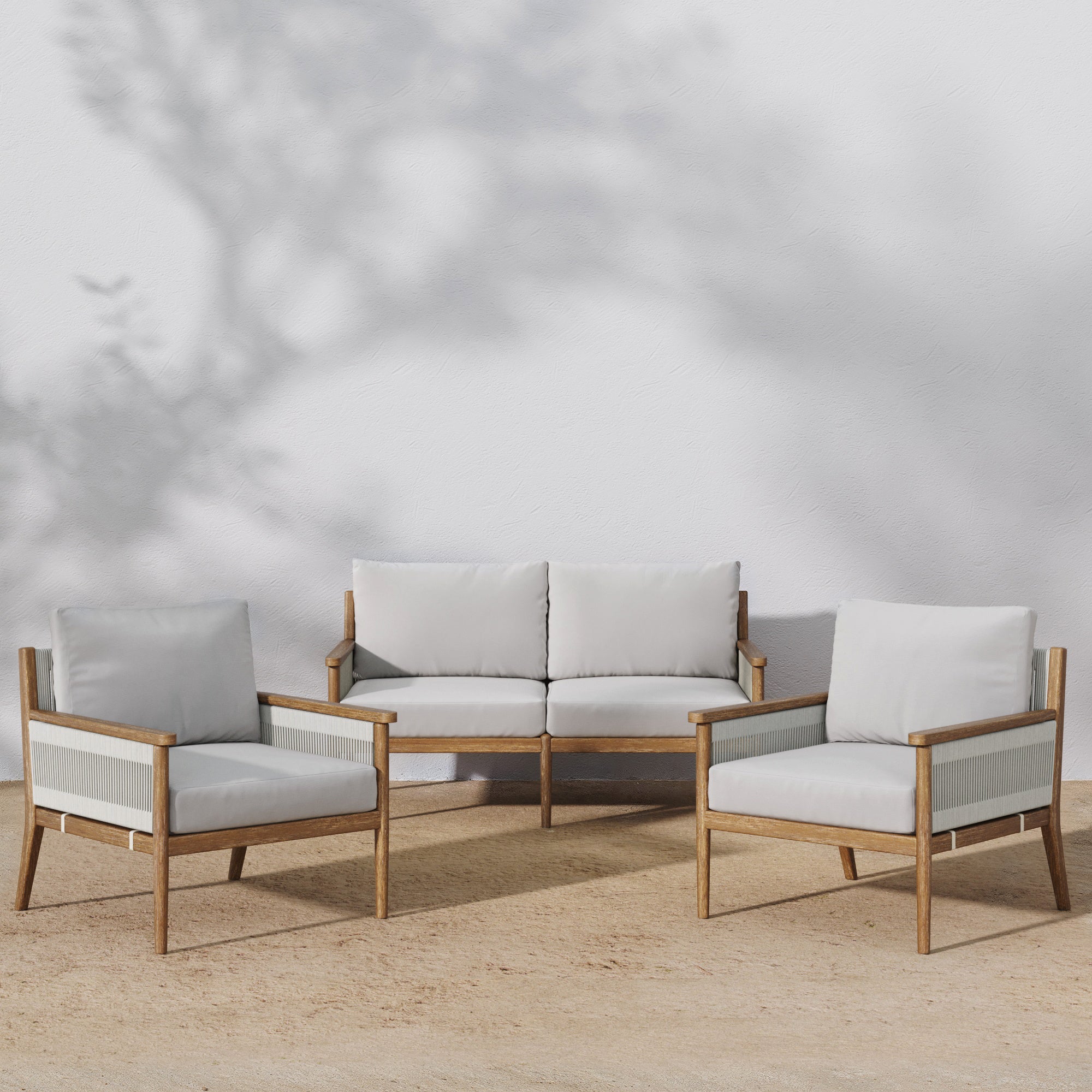 Three pieces of outdoor furniture—a two-seat sofa and two armchairs—feature light wood frames and white cushions, arranged on a sandy surface with soft shadows on a white wall behind them.