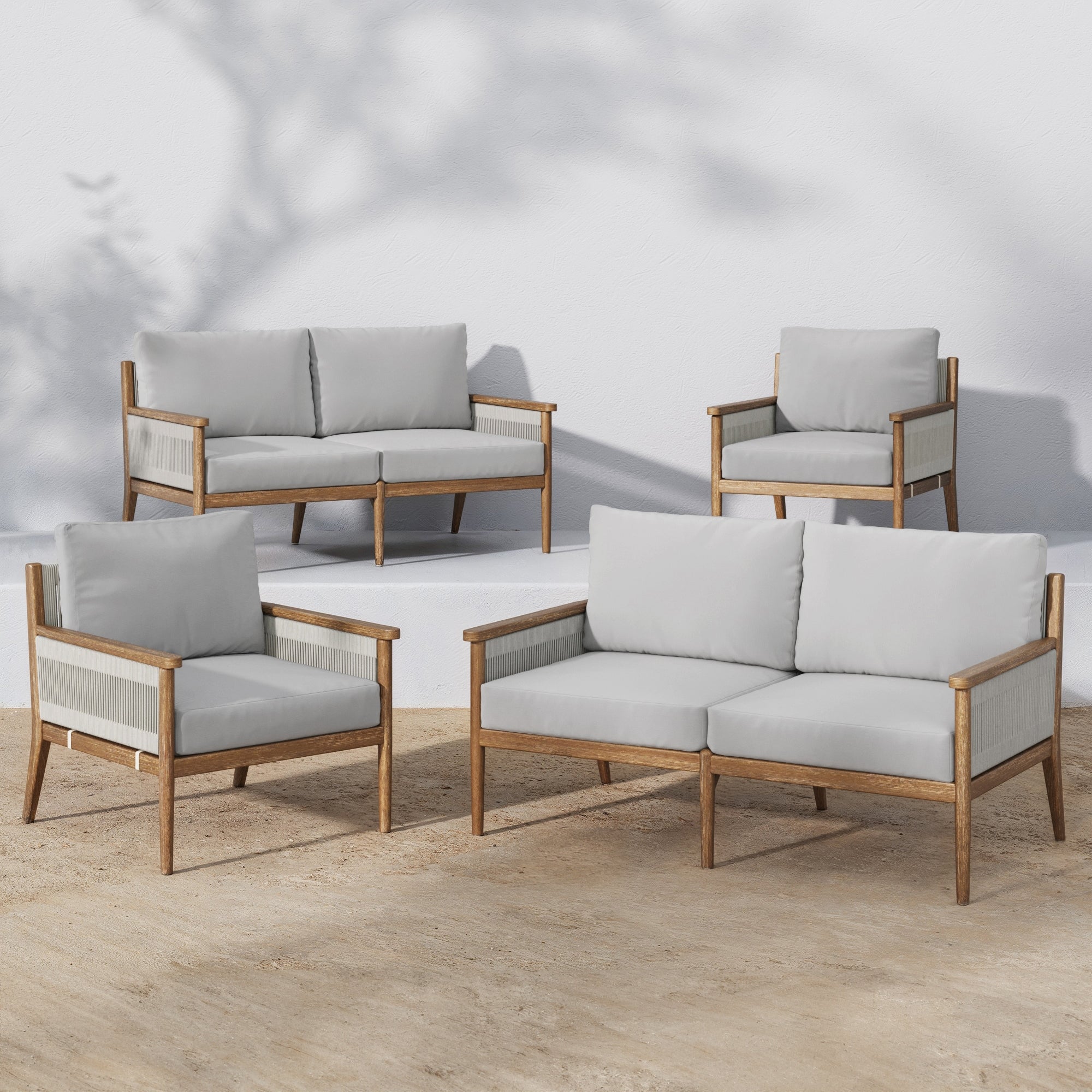 The Nathan James Outdoor 4 Piece Wood Patio Set features two loveseats and two armchairs with weatherproof acacia wood frames and light gray washable cushion covers, arranged on sand beside a white wall with tree branch shadows.