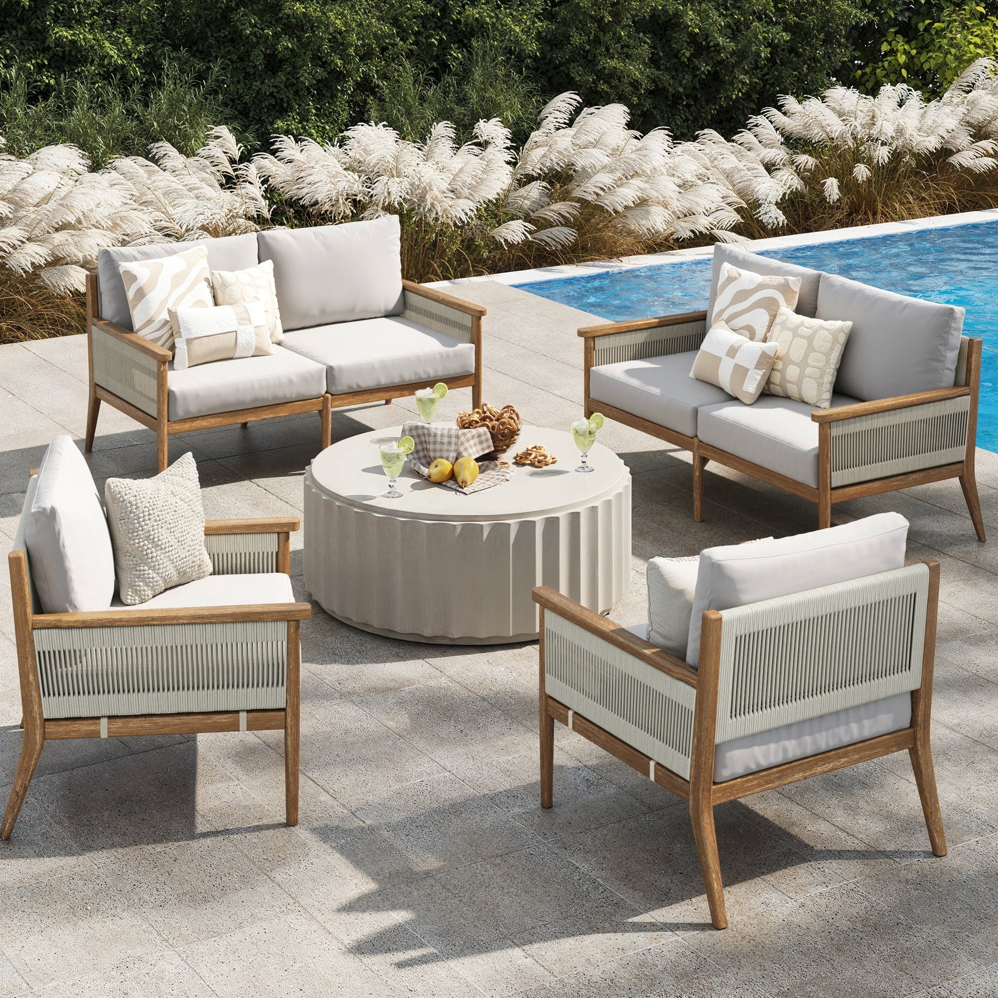 Outdoor patio seating area with four wooden chairs and a loveseat arranged around a round coffee table. Neutral cushions and patterned pillows, set beside a pool with green shrubs and ornamental grass in the background.