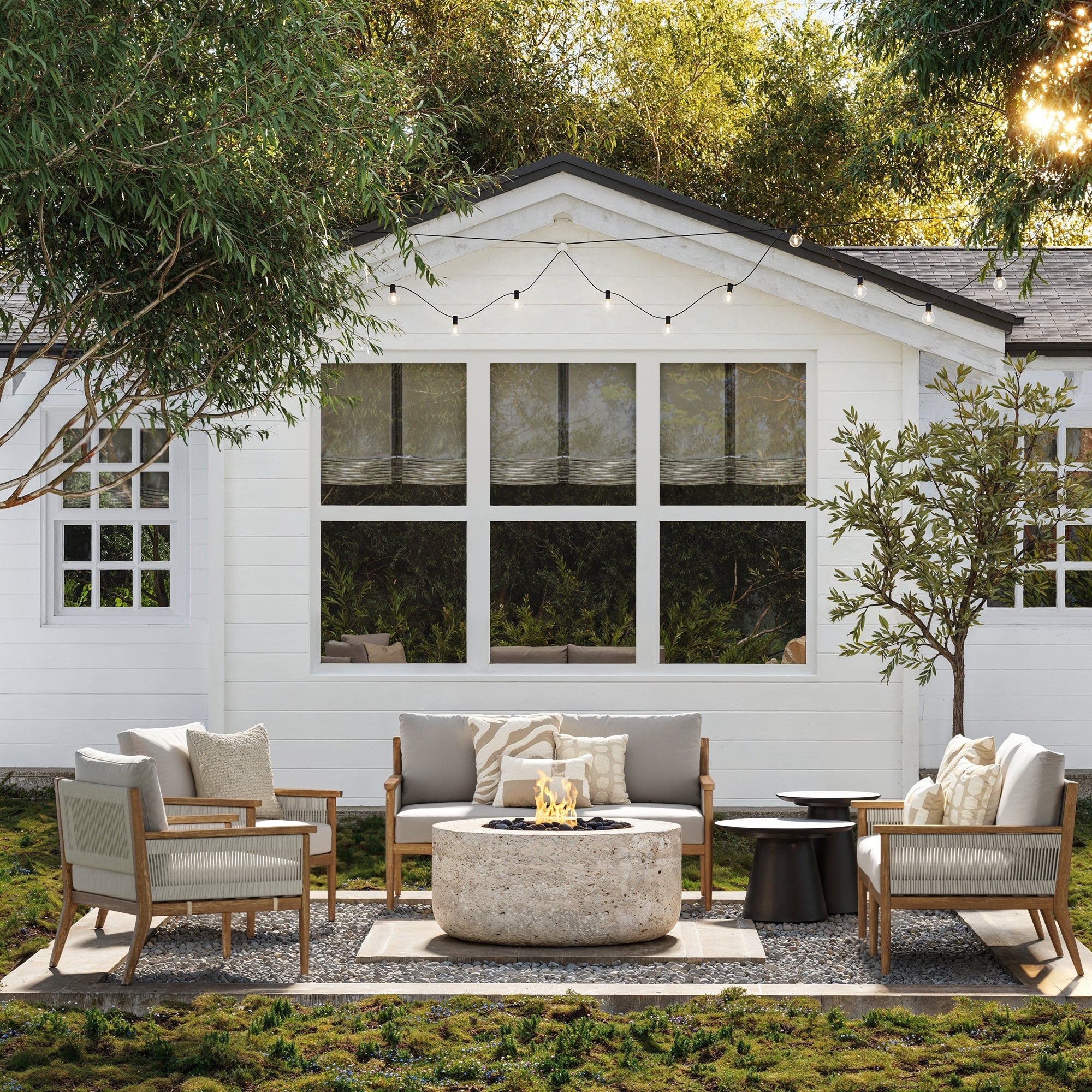 A welcoming patio showcases the Nathan James Outdoor 4 Piece Wood Patio Set Loveseats and Chairs with neutral, washable cushions, arranged around a round stone fire pit on gravel in front of a white house with string lights and trees.