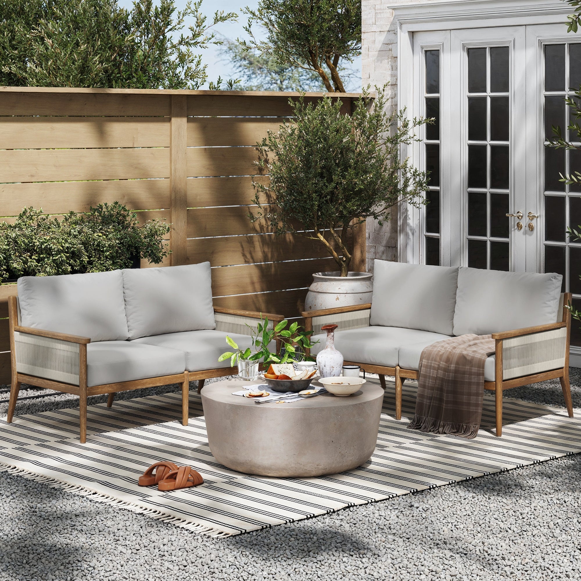 Outdoor patio setting with two cushioned wooden sofas, a round coffee table with drinks and snacks, a striped rug, potted plants, sandals, and a brown throw blanket, set against a wooden fence and glass doors.