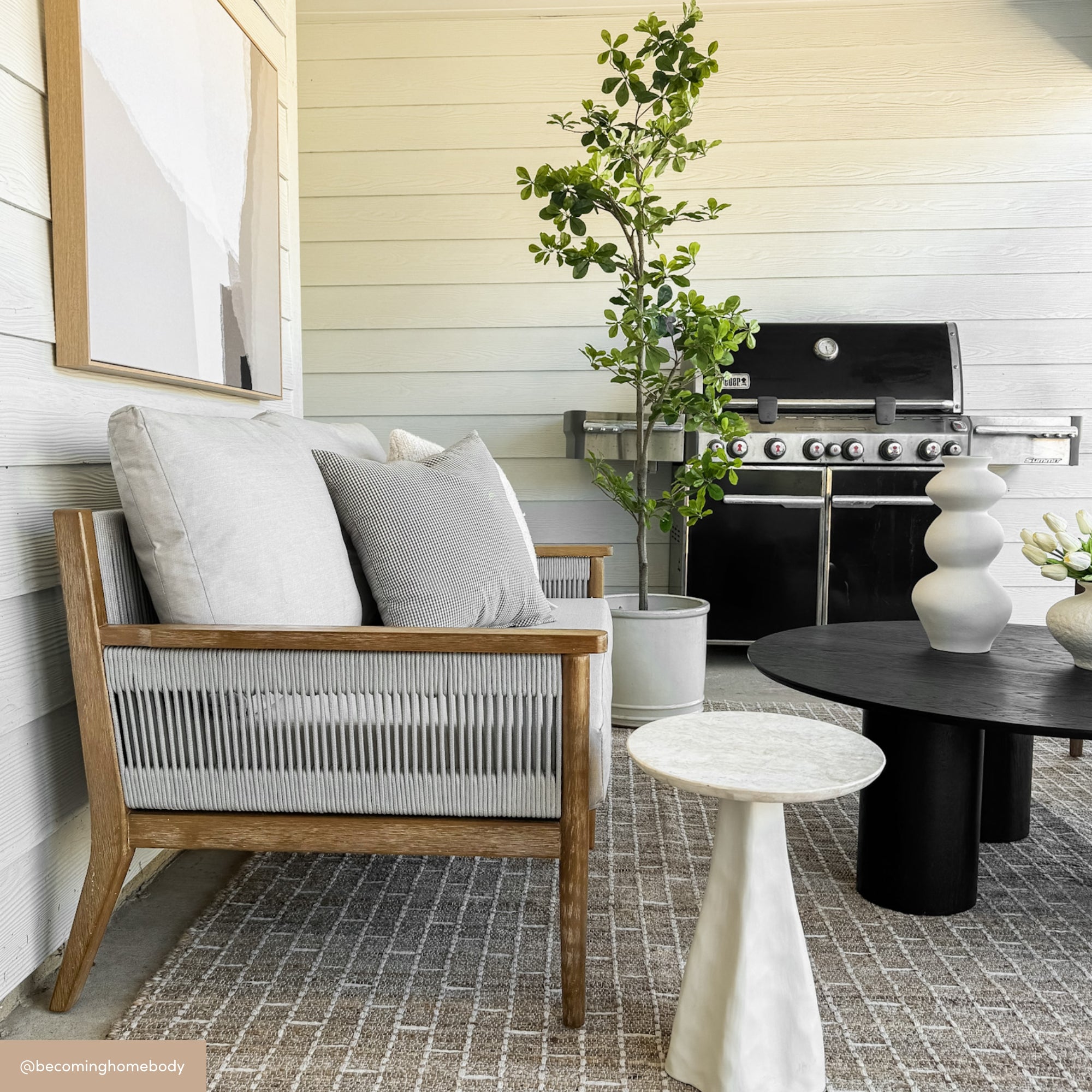 A modern patio featuring Nathan James Outdoor Wood & Rope Patio Set of 2 Loveseats in light gray, complemented by neutral decor, a small marble side table, large black grill, potted plant, and round black coffee table on a patterned rug.